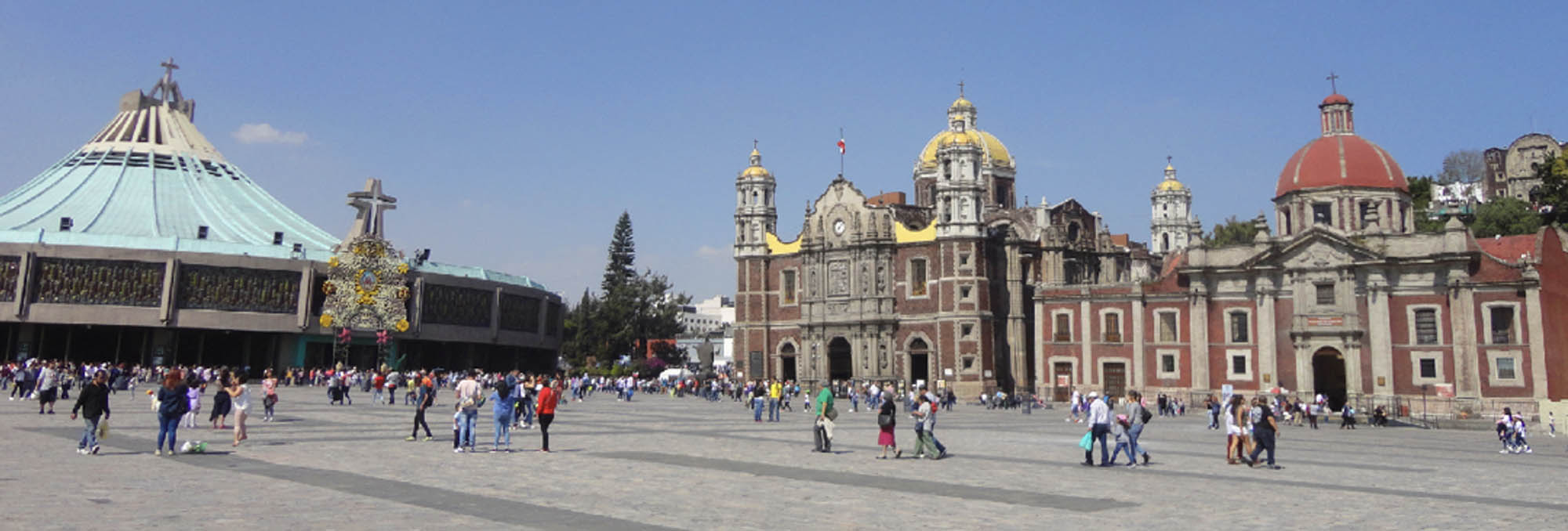 Photo of the Basilica with its large open plaza and numerous people walking around on a clear day. There are two main structures: one is circular and tent - like and the other features a red - domed roof and ornate facade.