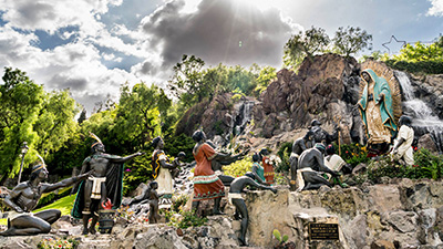 A group of statues on the Tepeyac Hill in Mexico City, the statues are positioned on a rocky terrain with a waterfall amongst a cloudy sky.