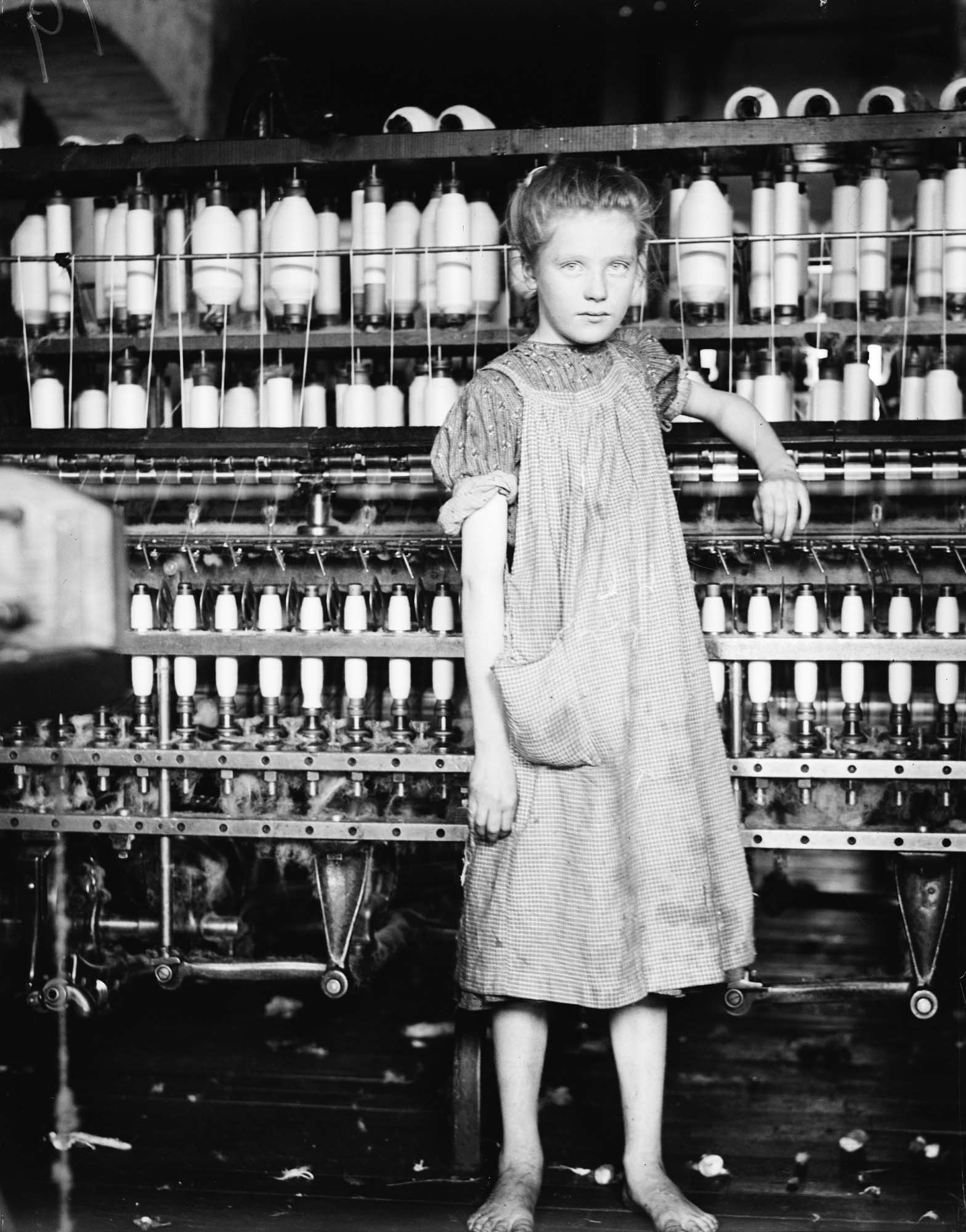 A young girl, wearing ragged clothing and without shoes, standing in front of a spinning machine at an industrial textile factory.