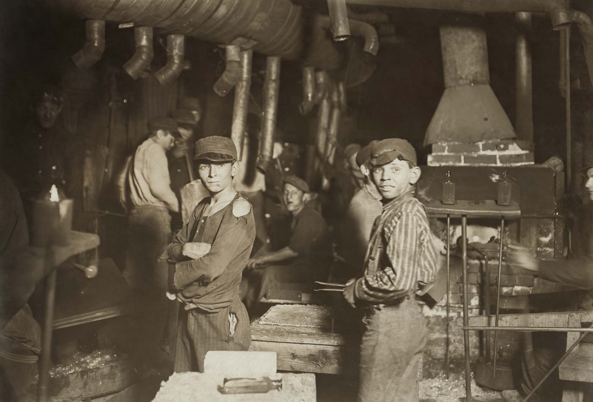 A photograph of young boys working with large machinery in a glass factory. The boys' faces are dirty and their clothing is ragged.