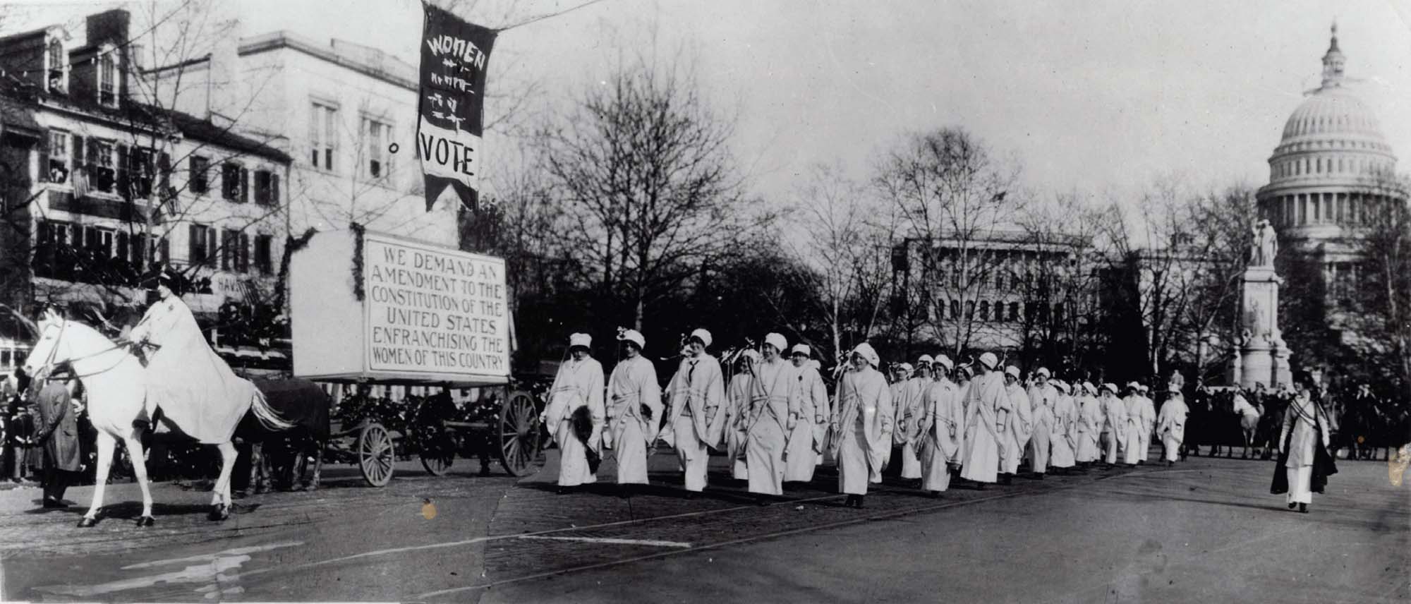 A parade of women, marching for the right to vote. Leading the march is a person on horseback, and behind them is a large sign demanding an amendment to the constitution to give women the right to vote.