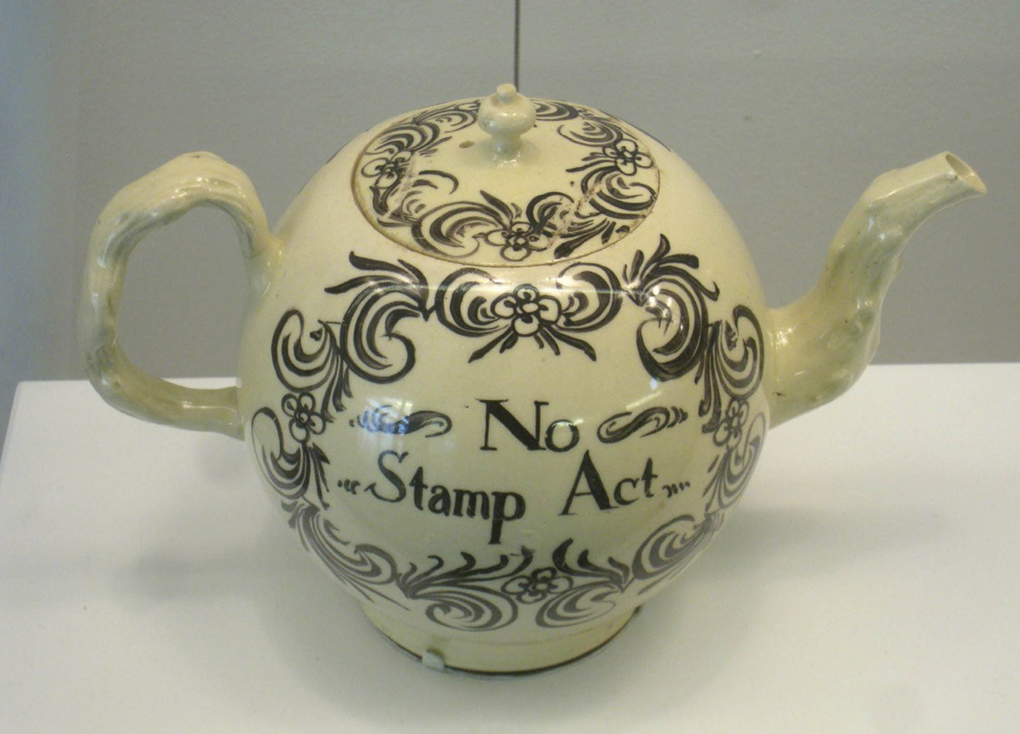 A teapot with a handle and lid, and the words “No Stamp Act” engraved in it.