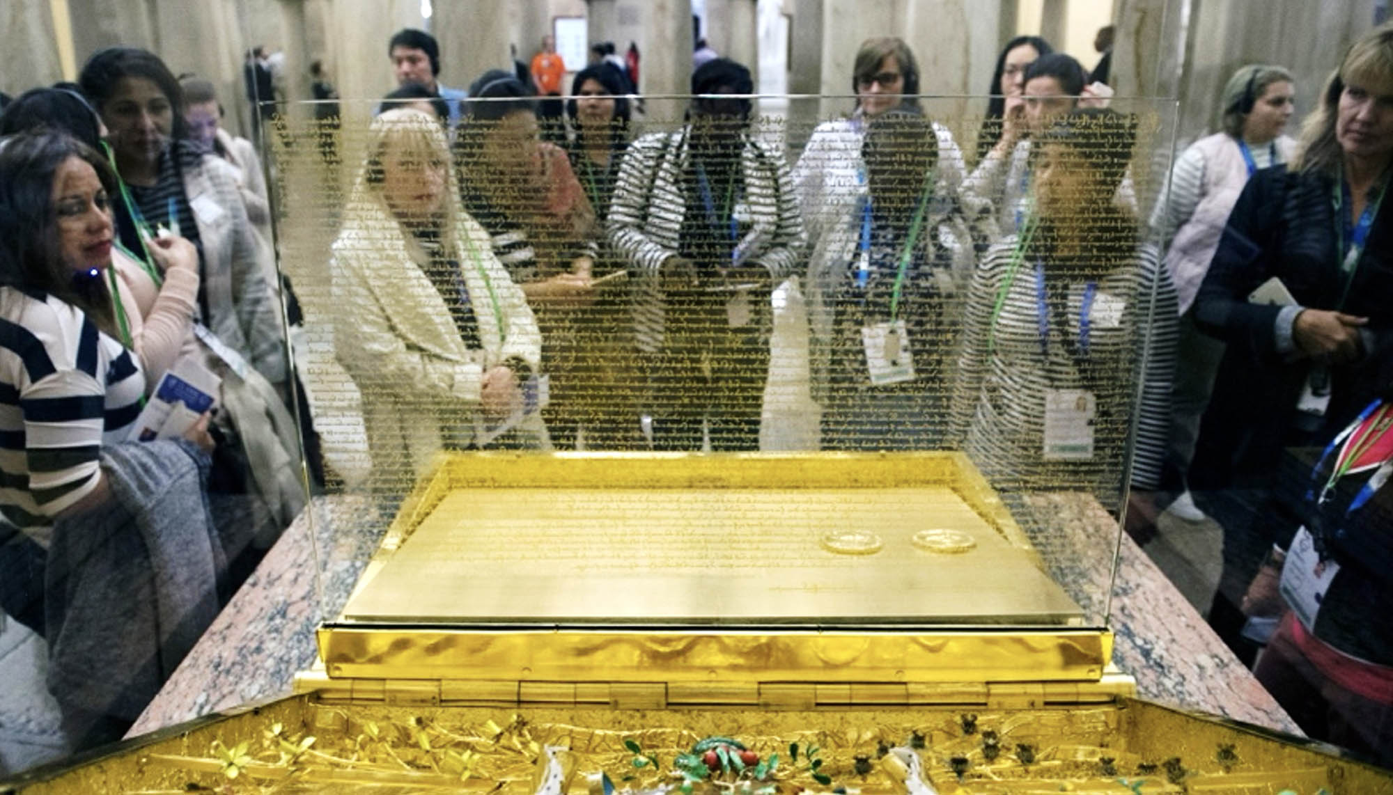A display case containing a document with two circular golden seals surrounded by a crowd of people, the document is displayed under protective glass and the case is ornate and golden.
