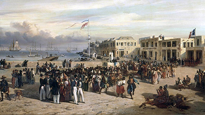 A large diverse crowd gathered on a coastal area with several ships visible on the sea in the background, a French flag is displayed on a pole.