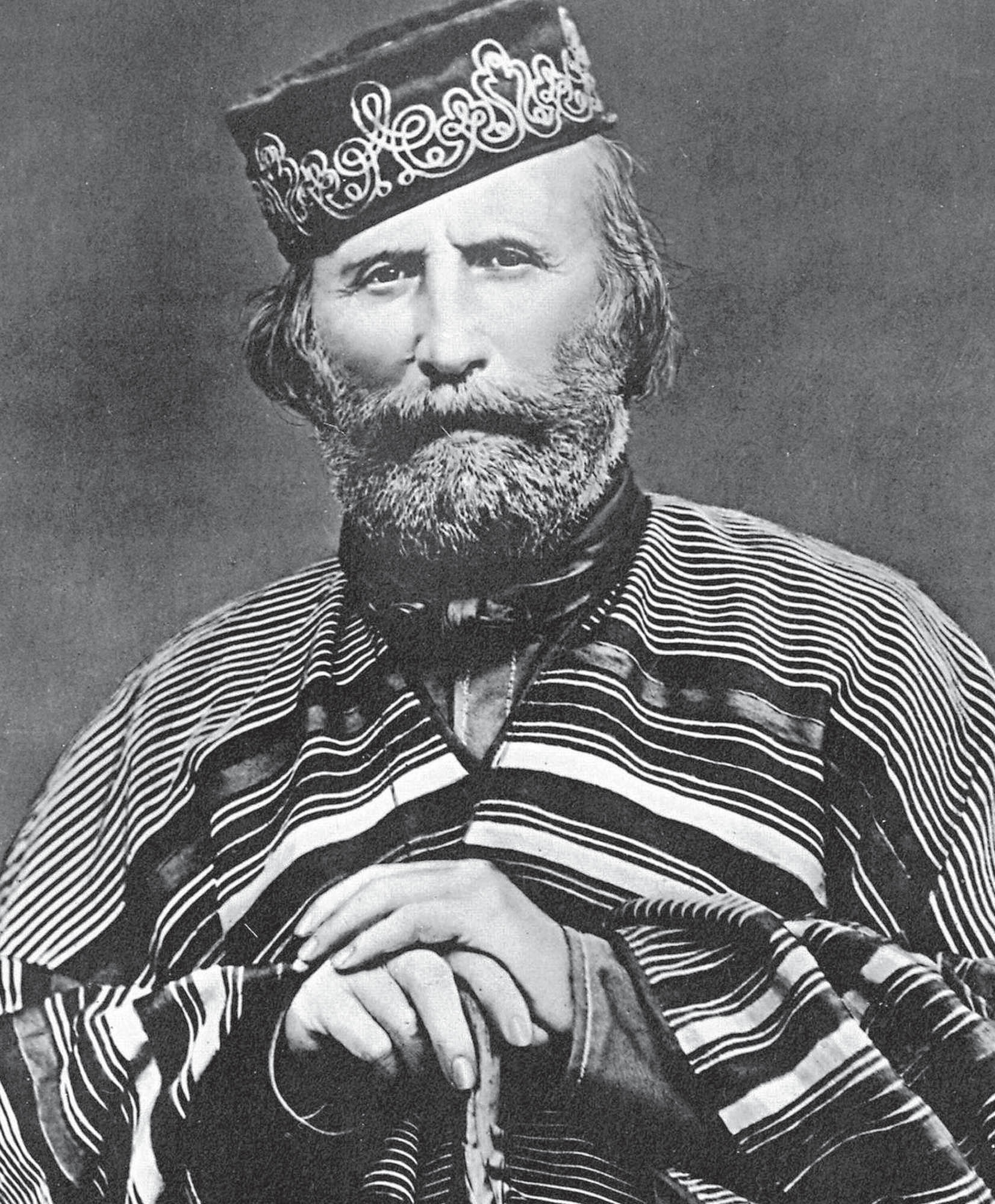 A black and white portrait of Giuseppe Garibaldi, he has a full beard and mustache, is wearing a decorative cap and striped poncho, and is holding a cane.