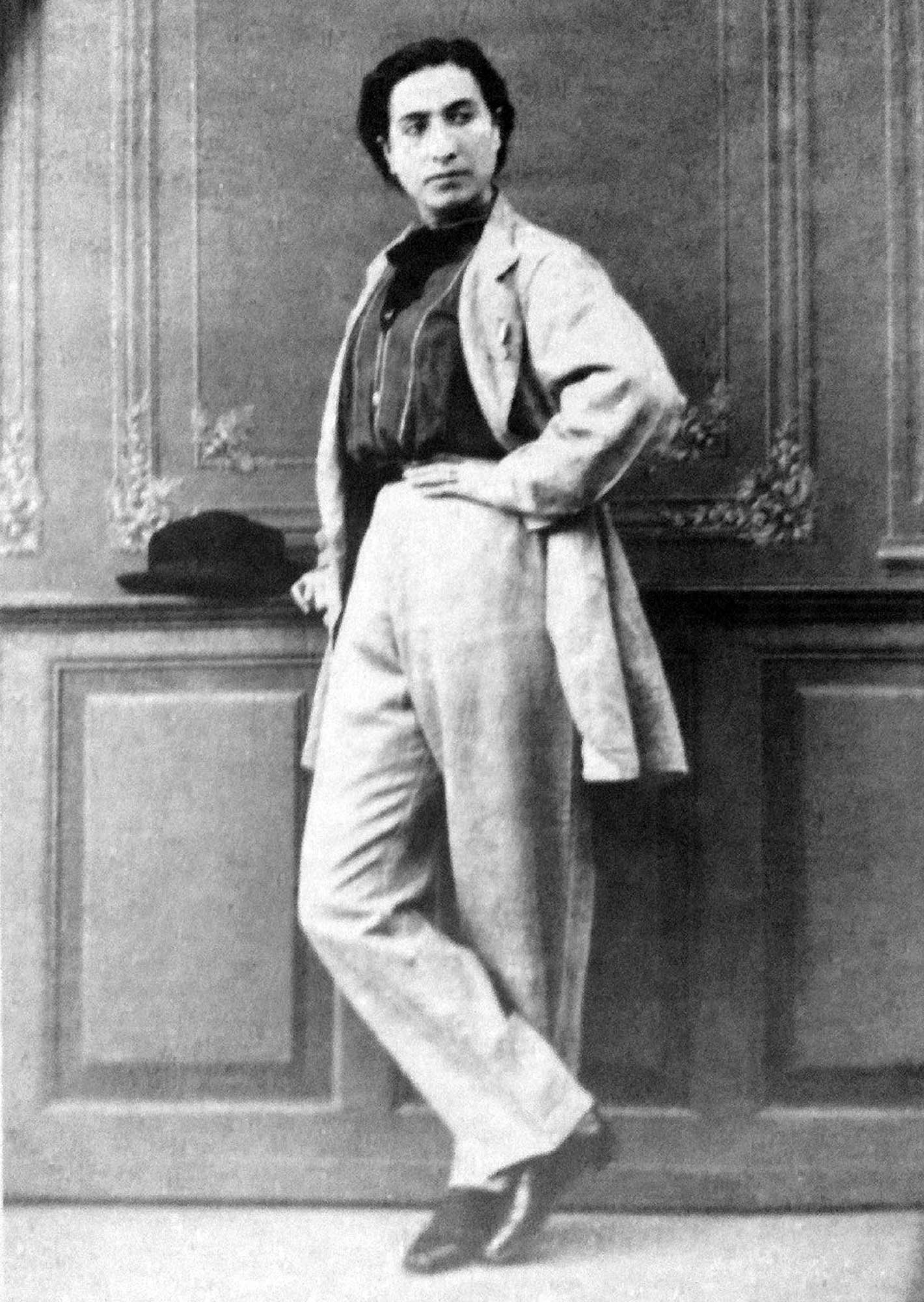 A black and white portrait of Anita, who is standing with one hand resting near a hat placed on a ledge and the other on their hip; they are looking off to the left with a serious expression.