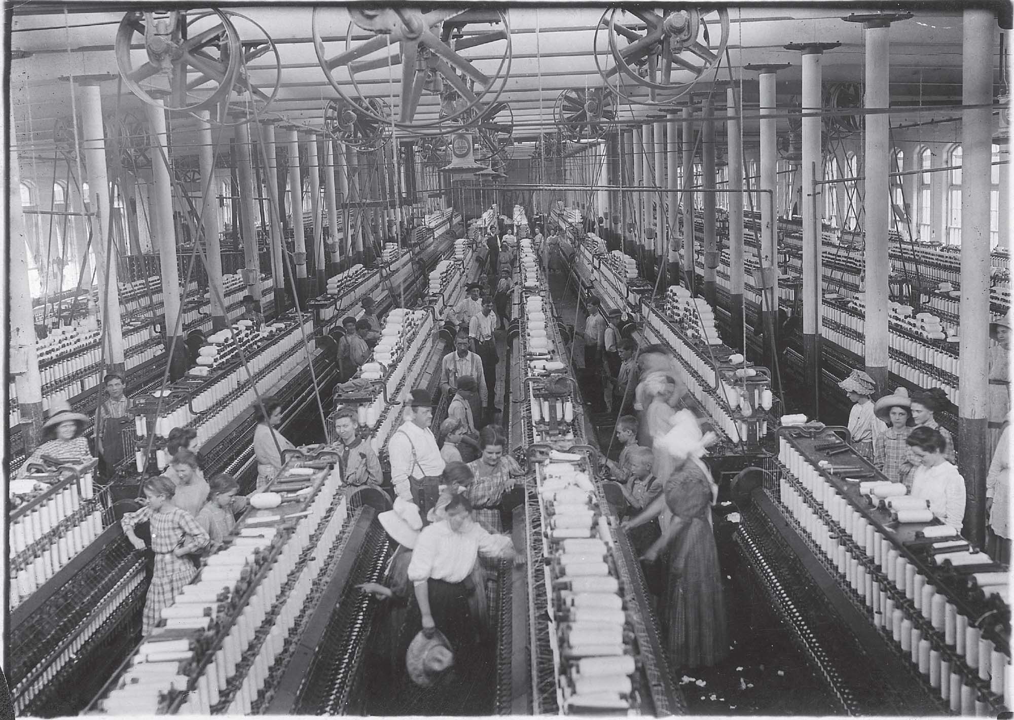 A black and white photograph of people working in a large, industrial factory. People are working at large, yarn spinning machines.