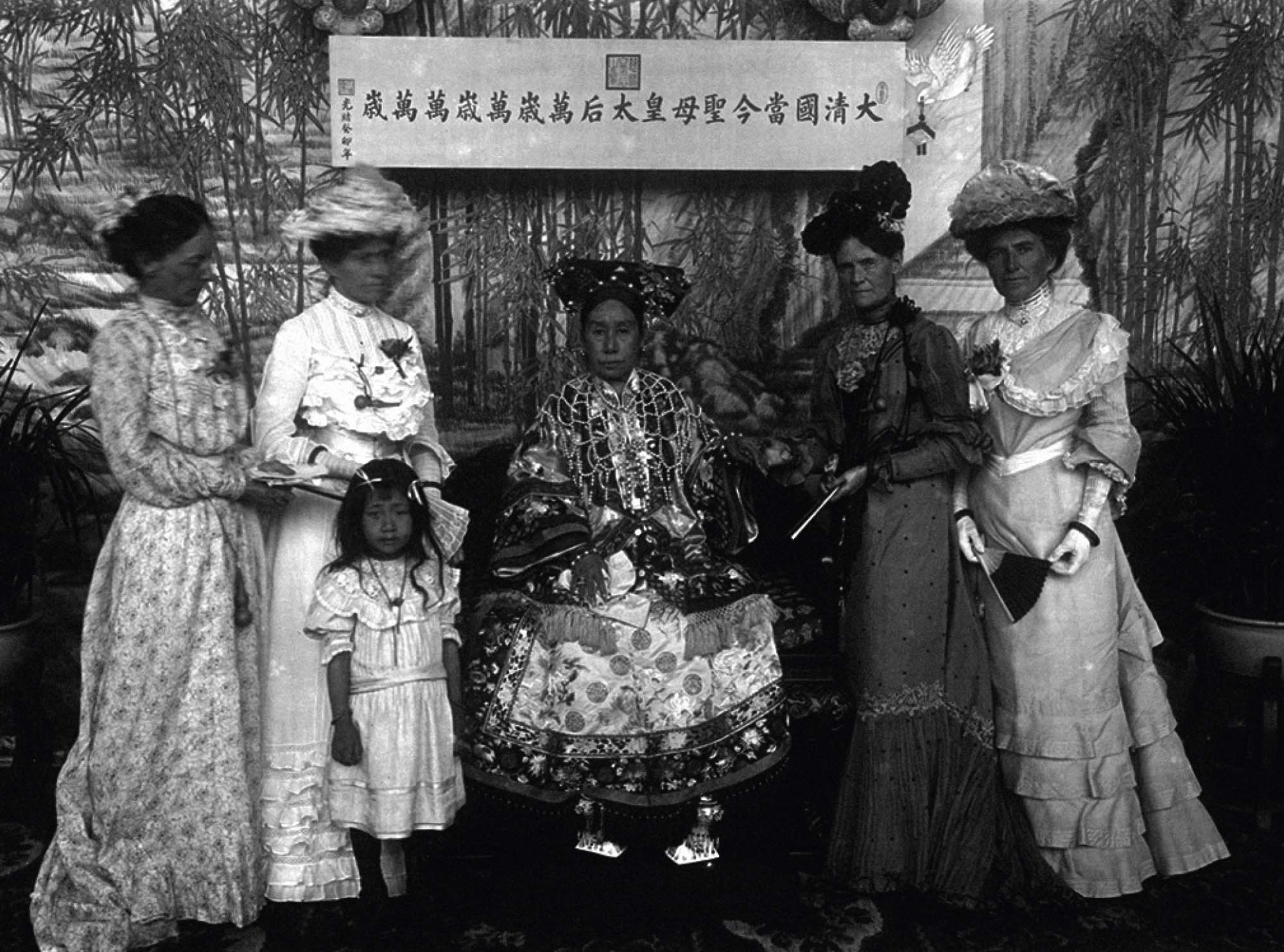 Photograph of the Empress Dowager Cixi, dressed in sparkling, ornate clothing, and seated in a chair. Surrounding her are four other women, standing, and a young girl.
