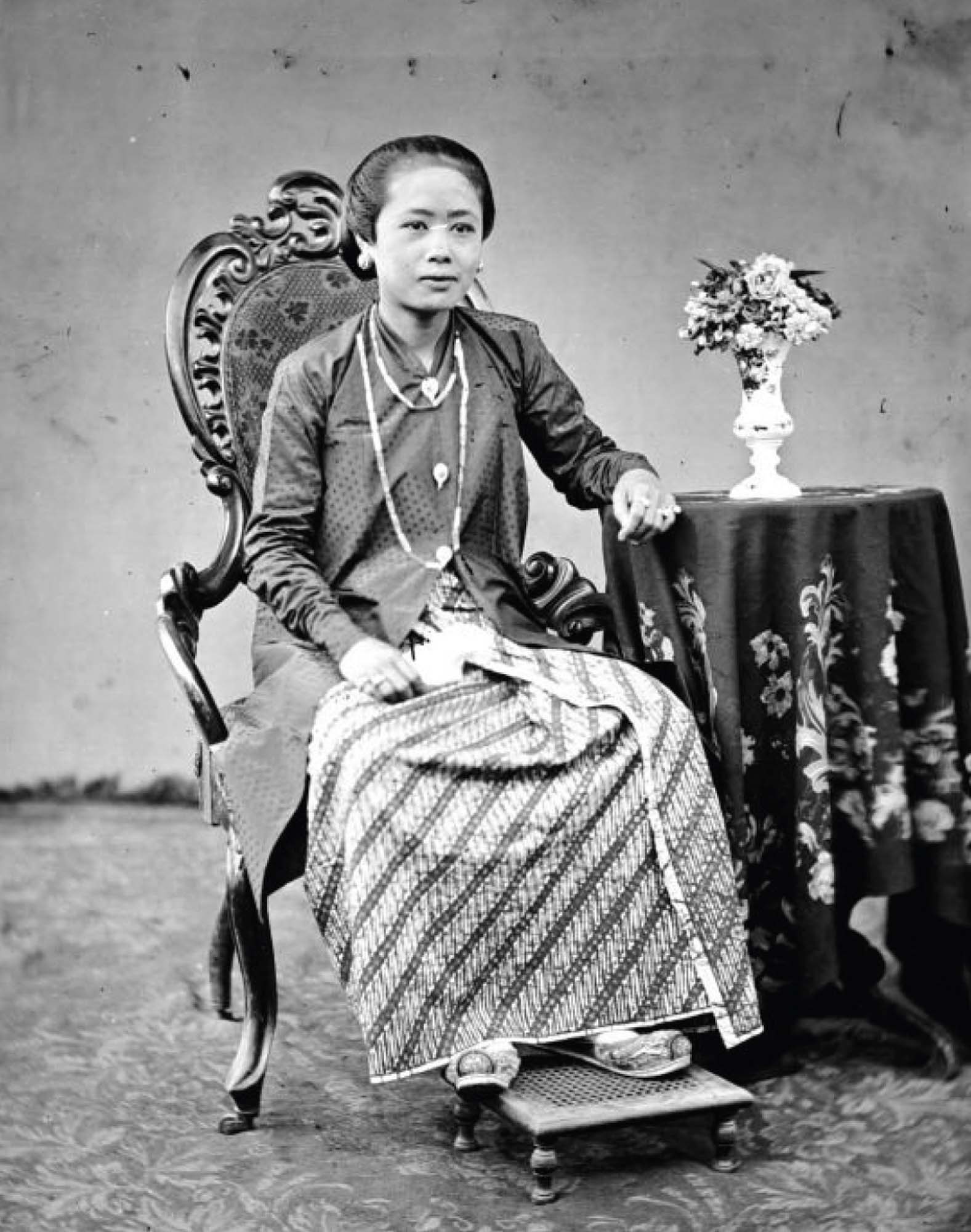 A photograph of a woman, seated, her feet resting on a small foot stool. Her arm is resting on a small, round table covered in a floral tablecloth.