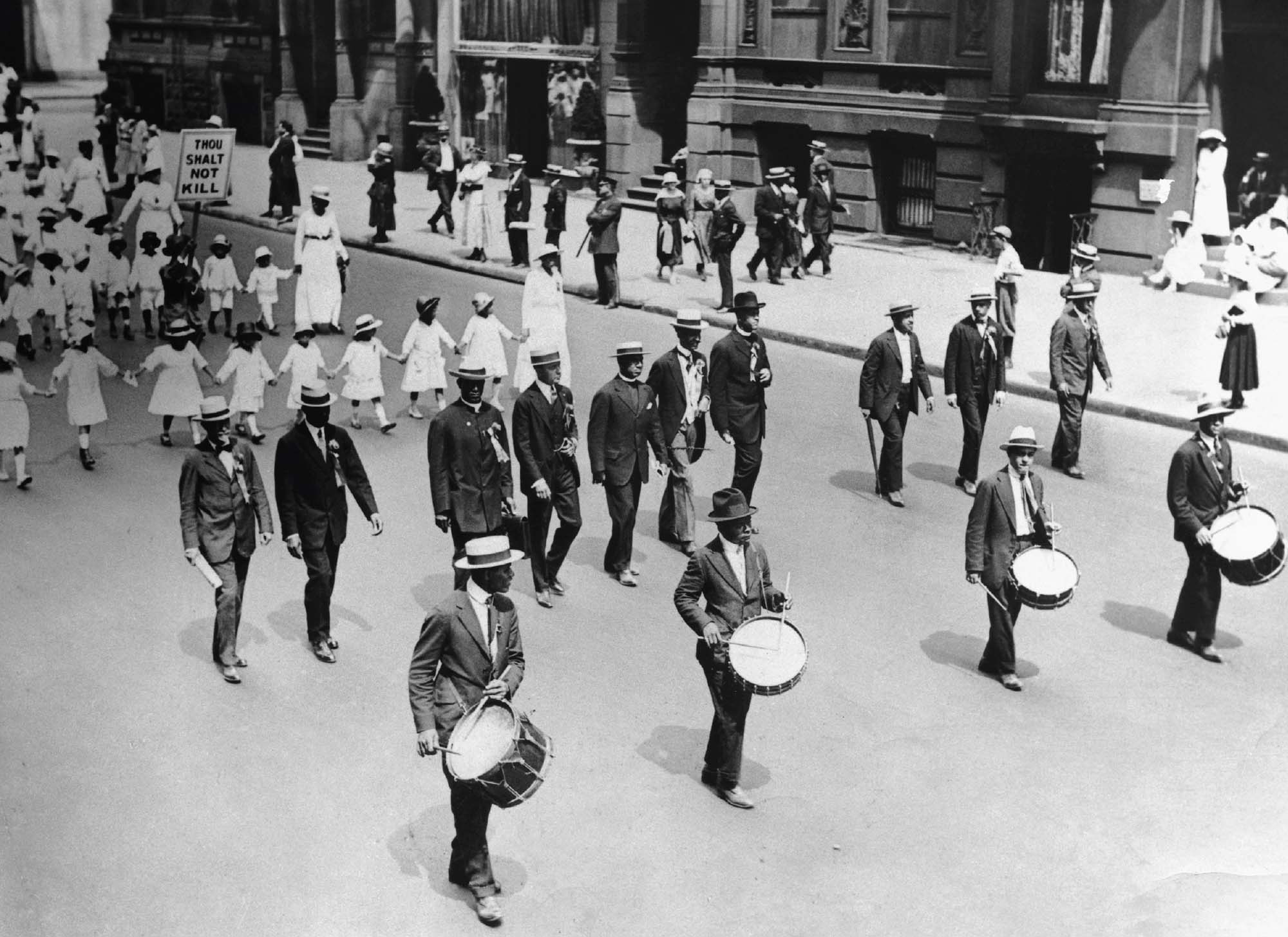 Black and white photograph of people marching in protest down the street. Four men in front carry drums, and behind them a line of children walk. One child holds up a sign that reads “Thou shalt not kill”.