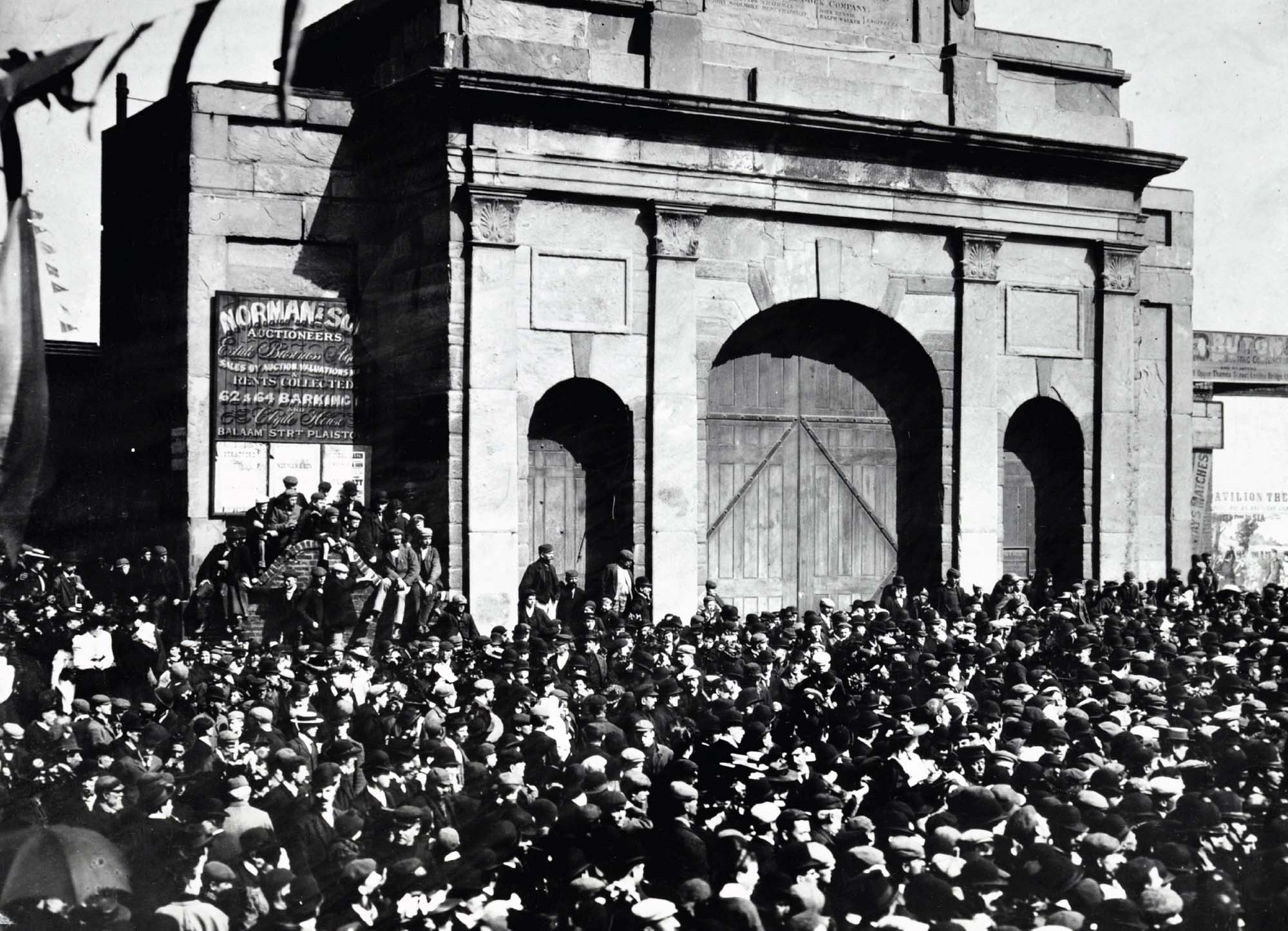 A black and white photograph of a vast amount of people standing outside of a large wooden gate.