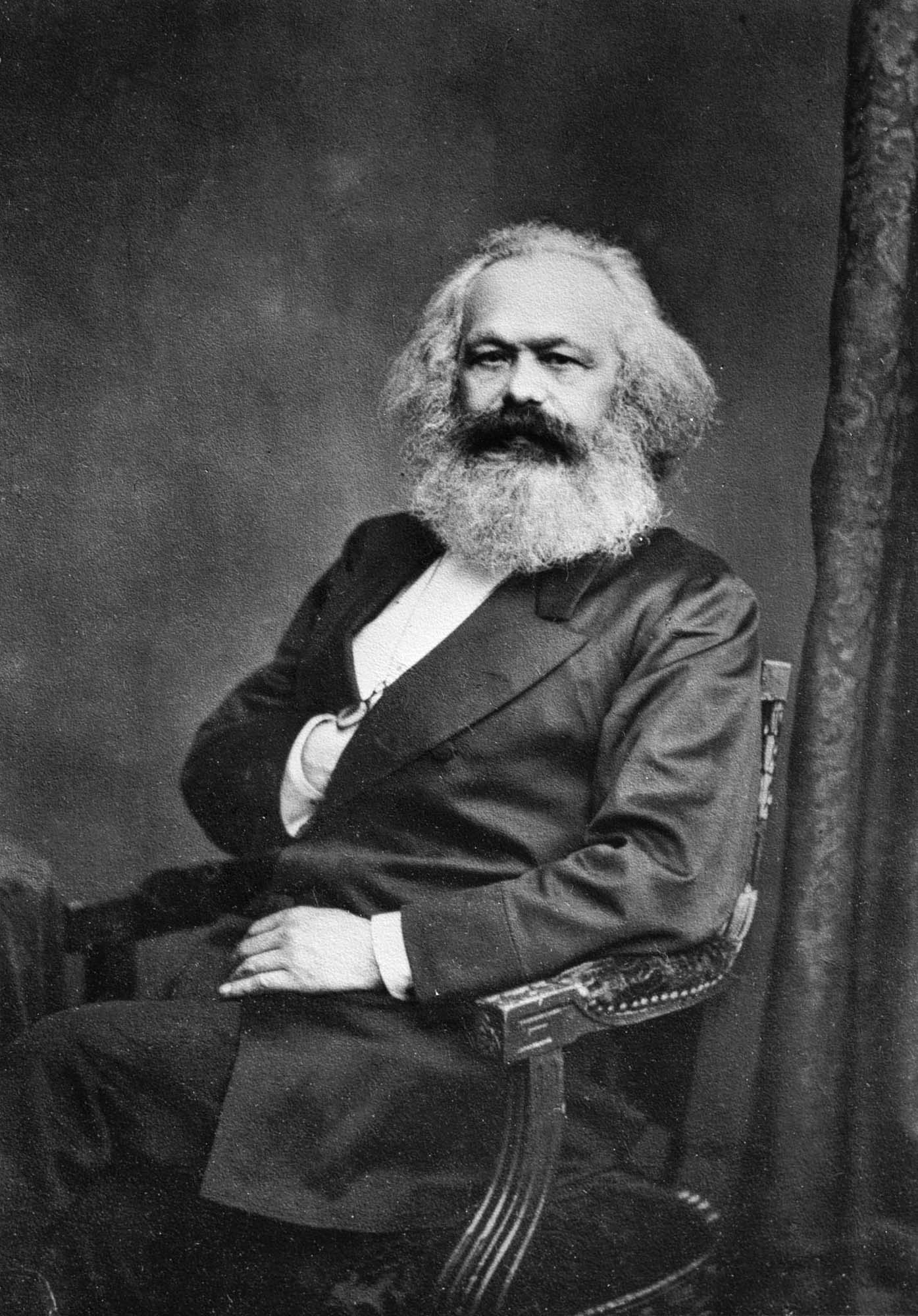 A portrait of German social scientist Karl Marx seated in a chair, with his right hand tucked into the suit jacket.