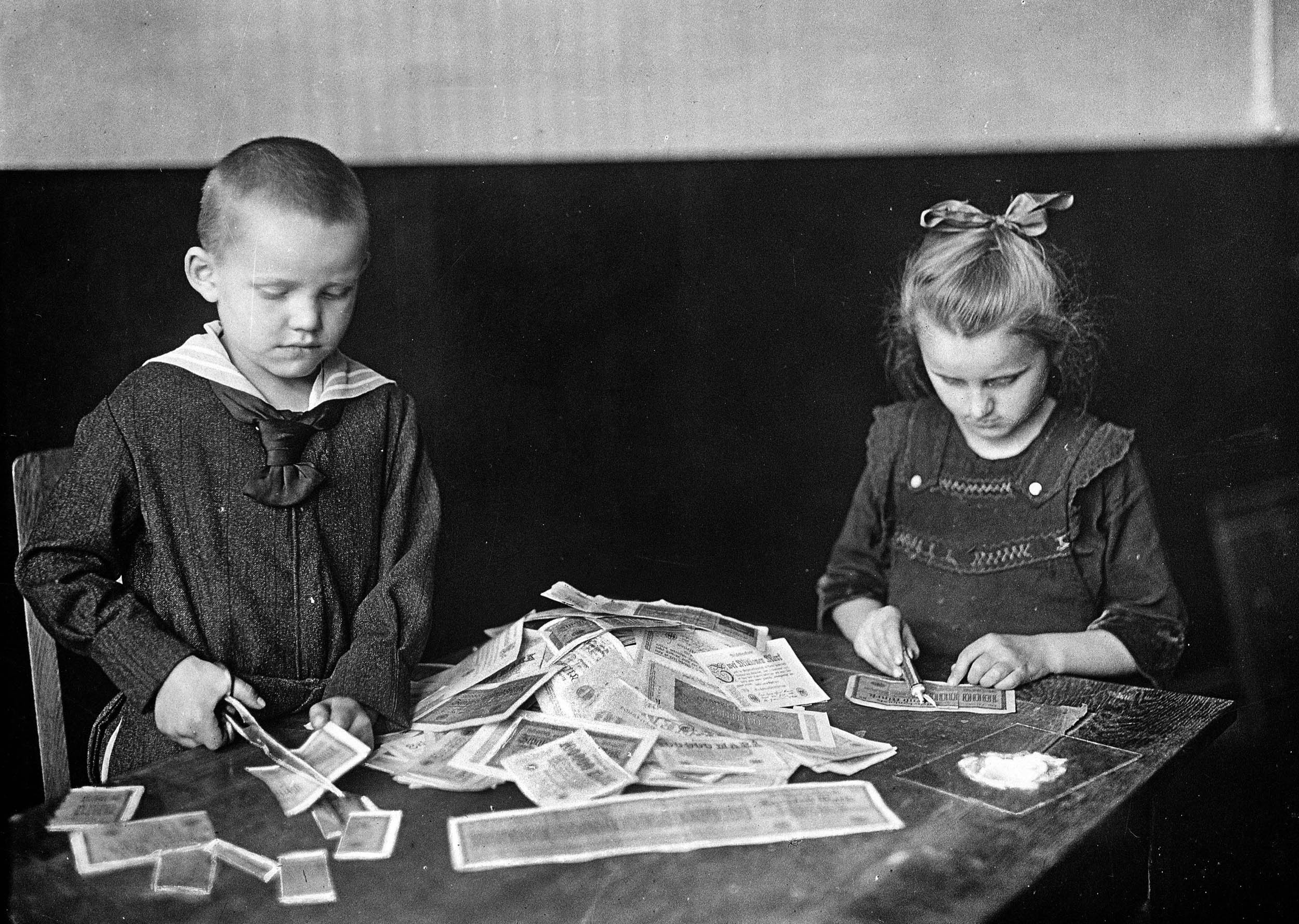 A black and white photo of two young children seated at a table, cutting paper banknotes into pieces, with a large pile of notes in front of them.