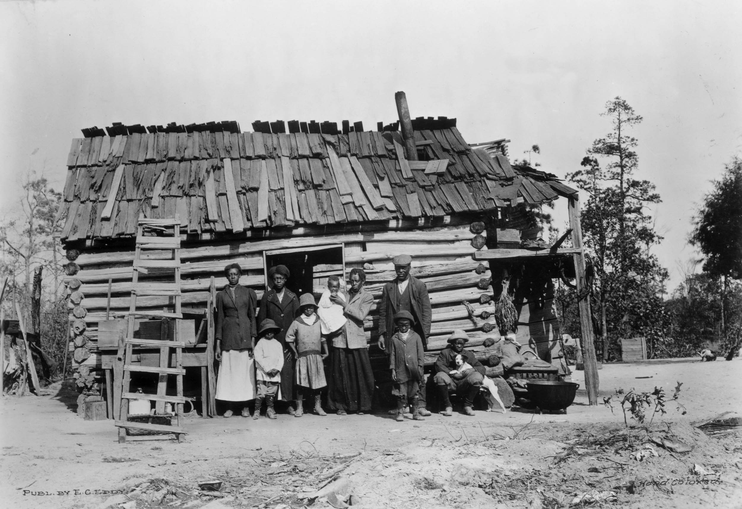 Black-and-white photo of an African American family standing outside a log cabin with a wooden shingle roof in the rural southern United States.