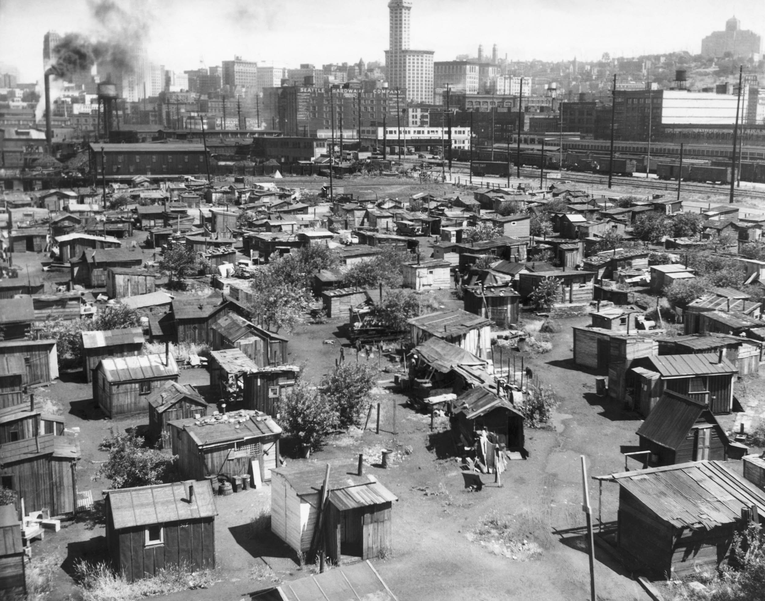 Black-and-white photo of a Depression-era shantytown with rows of makeshift wooden shelters in front of an industrial cityscape.