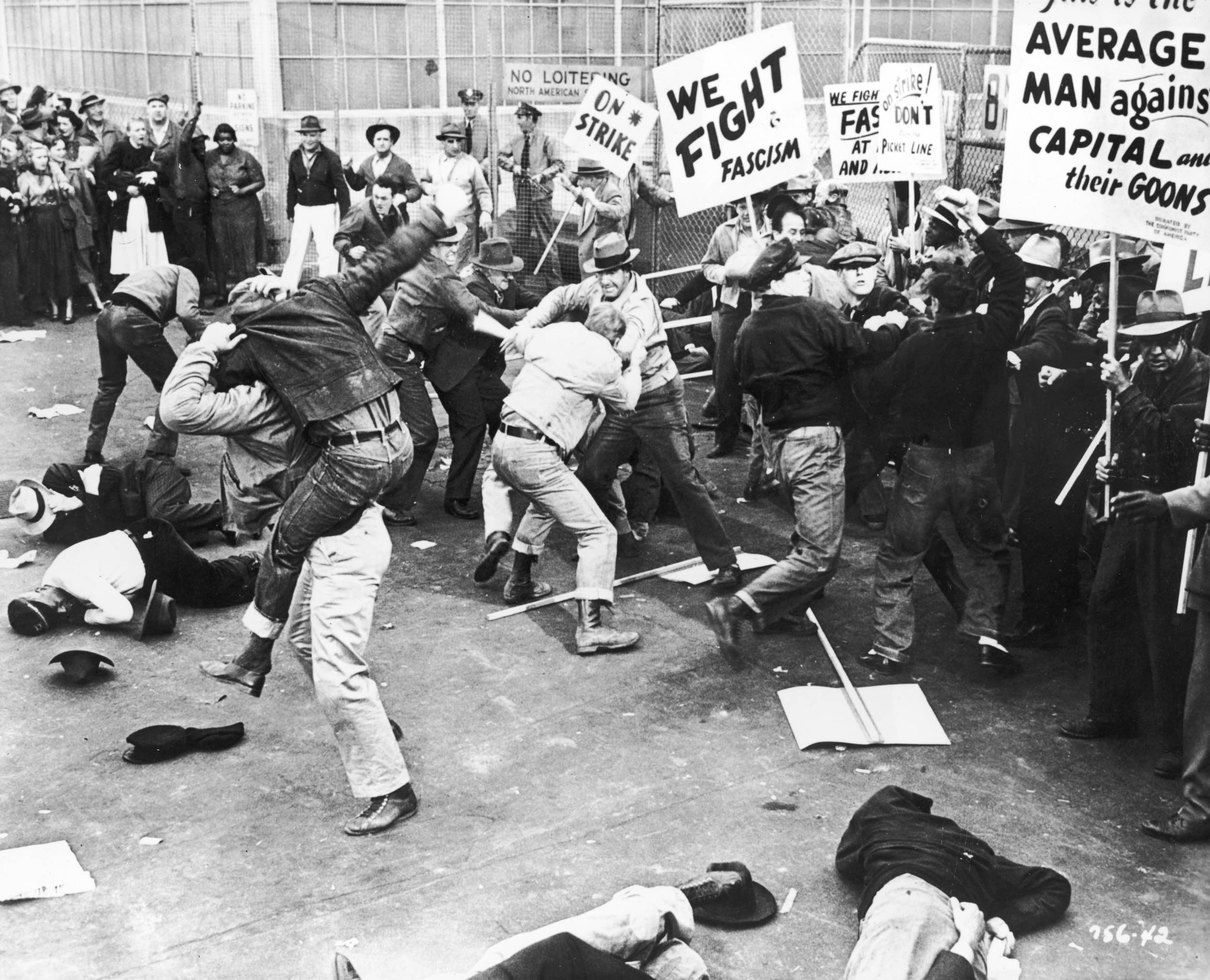 Black-and-white photo of a violent clash between striking workers and police or strike-breakers, with protest signs reading “We Fight Fascism” and “On Strike” amid the chaos.