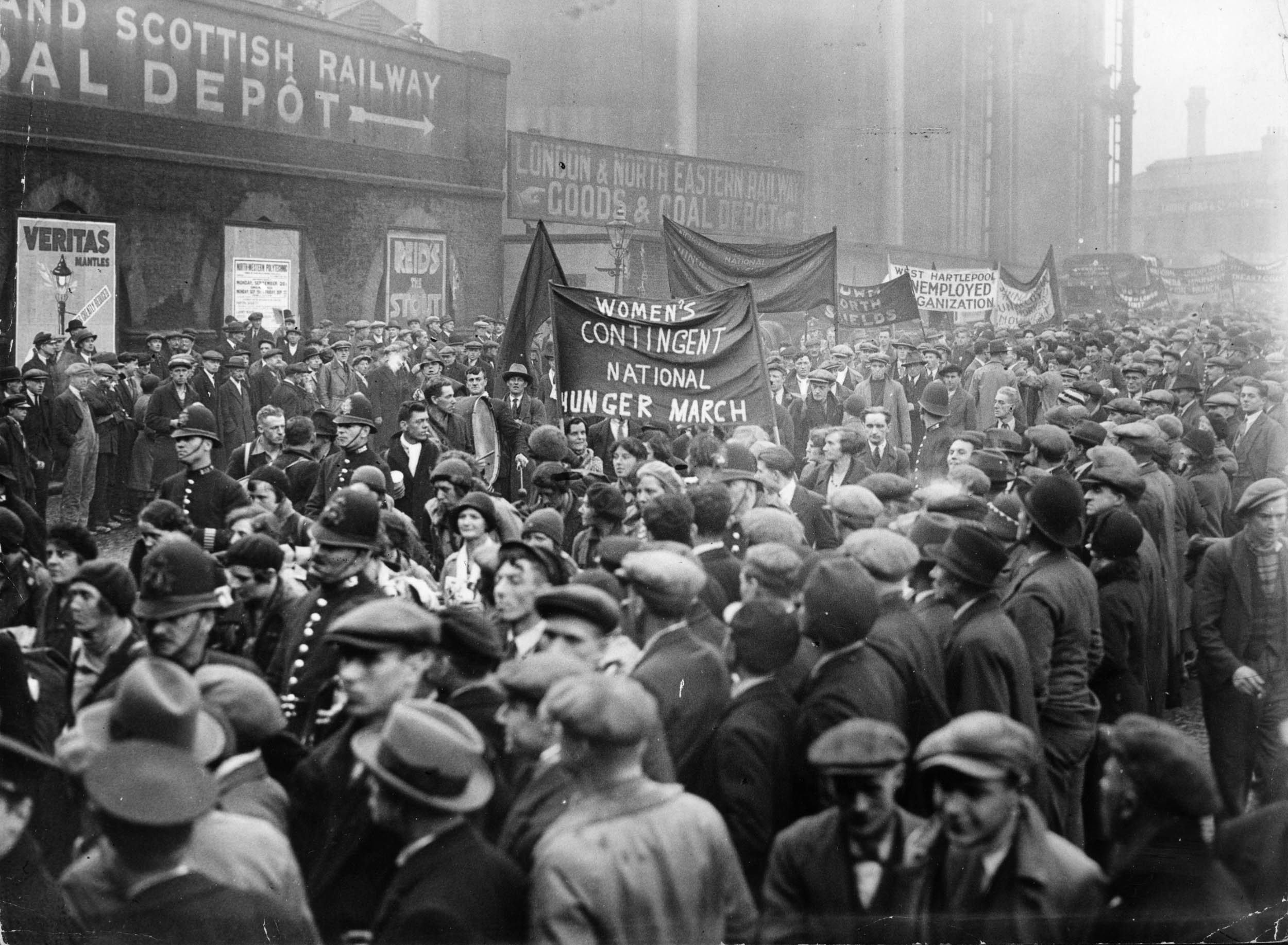 Black-and-white photo of a dense crowd of men and women in coats and flat caps, many facing forward, with police officers lining the sides and people holding large cloth banners in the background.