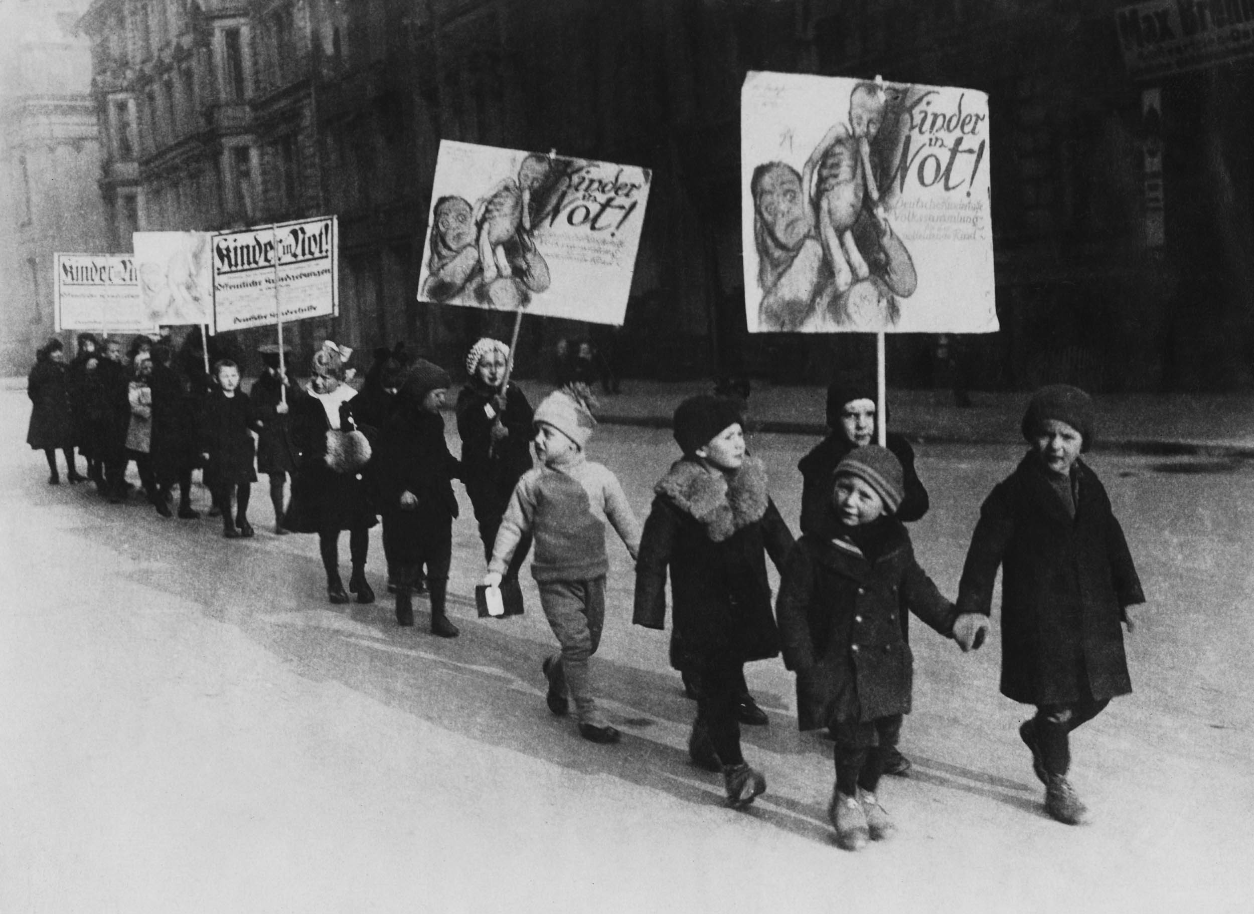 Black-and-white photo of children walking in a line down a city street, bundled in winter coats and hats, holding large, illustrated protest signs.
