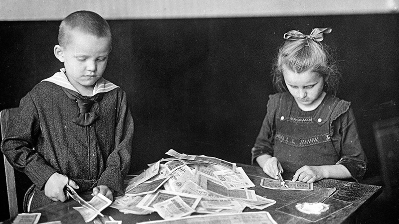 A black and white photo of two young children seated at a table, cutting paper banknotes into pieces, with a large pile of notes in front of them.