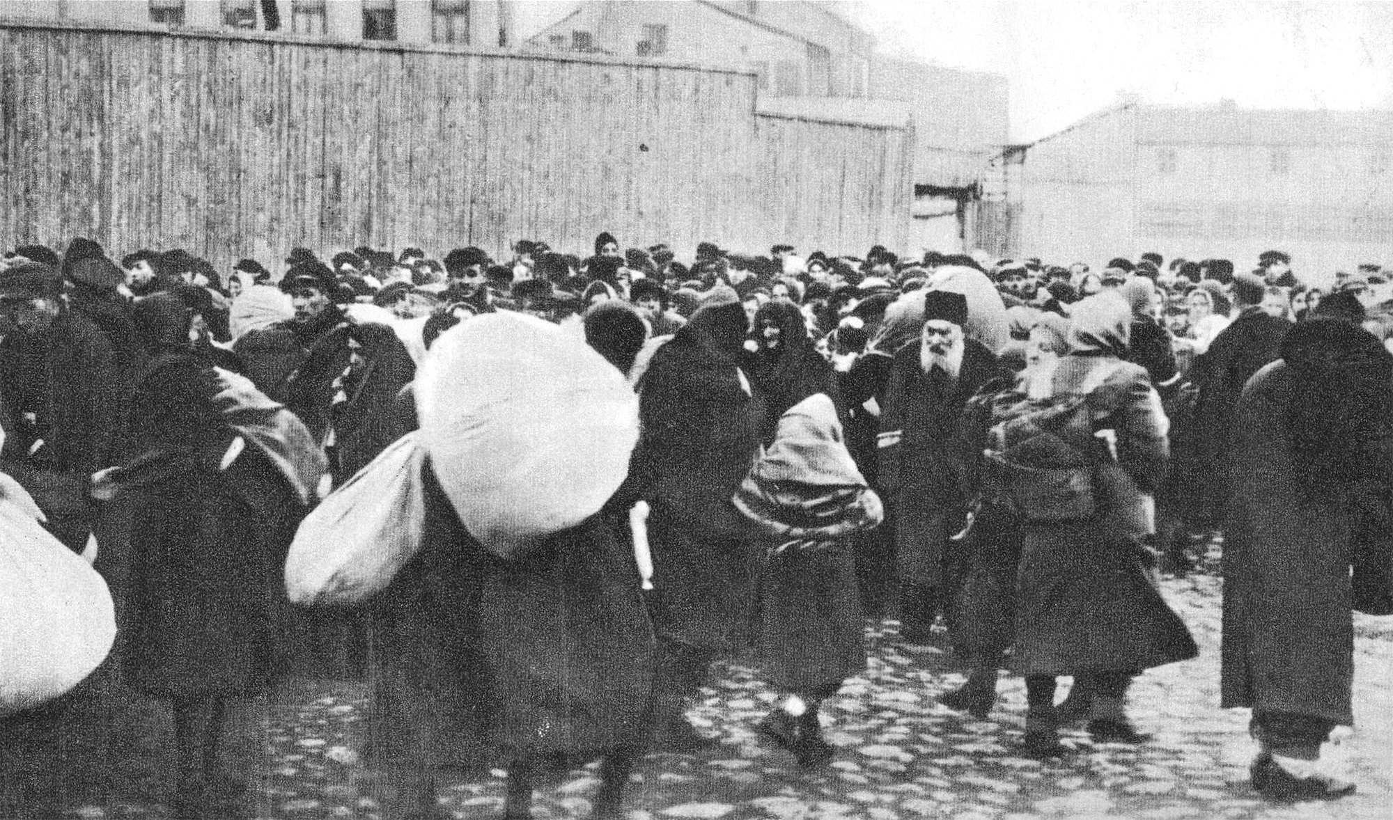 A black - and - white photograph of a crowded street scene with many people wearing coats and carrying large bundles, gathered closely together near wooden fences and buildings.