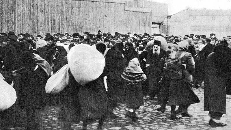 A black-and-white photograph of a crowded street scene with many people wearing coats and carrying large bundles, gathered closely together near wooden fences and buildings.