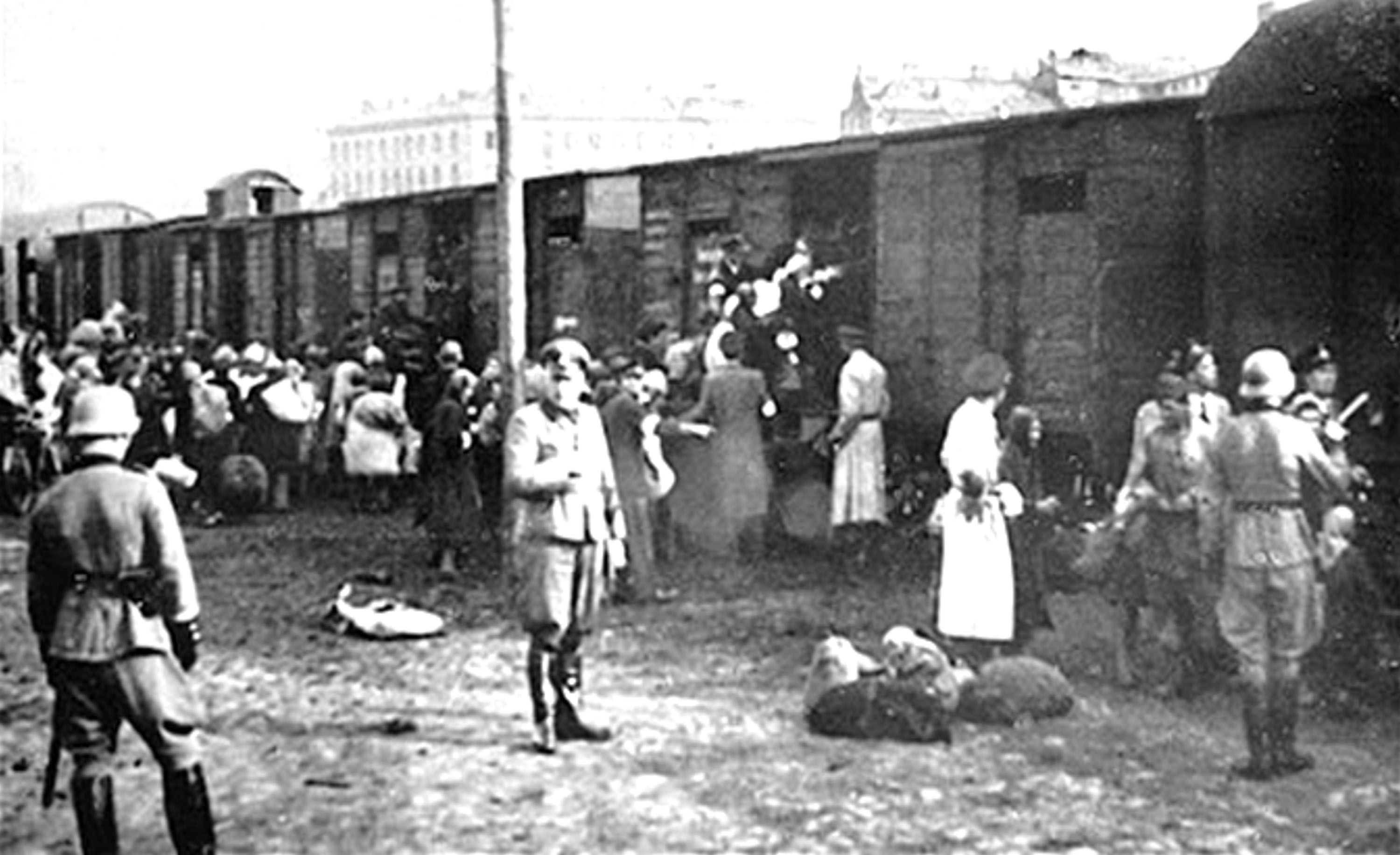 A black-and-white photograph showing groups of people gathered beside a long row of train cars, with uniformed guards standing nearby and some individuals carrying bundles.