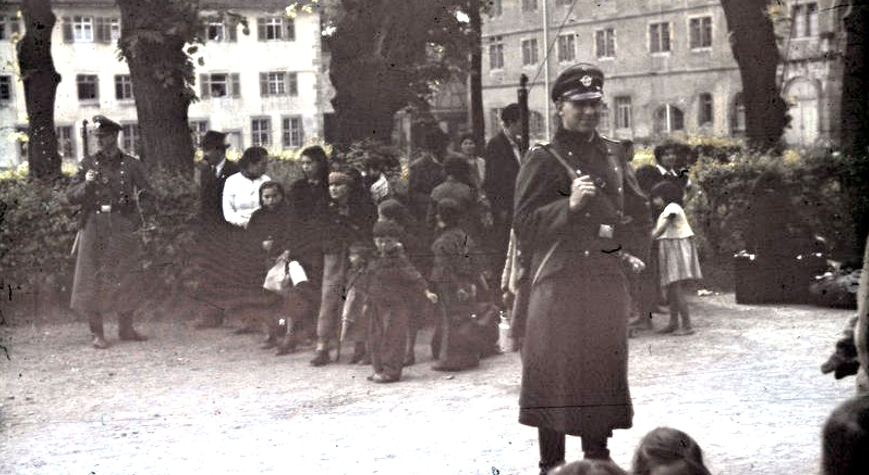 A black and white photograph showing a group of people, including children, gathered outdoors near buildings, with uniformed guards standing in the foreground and background.