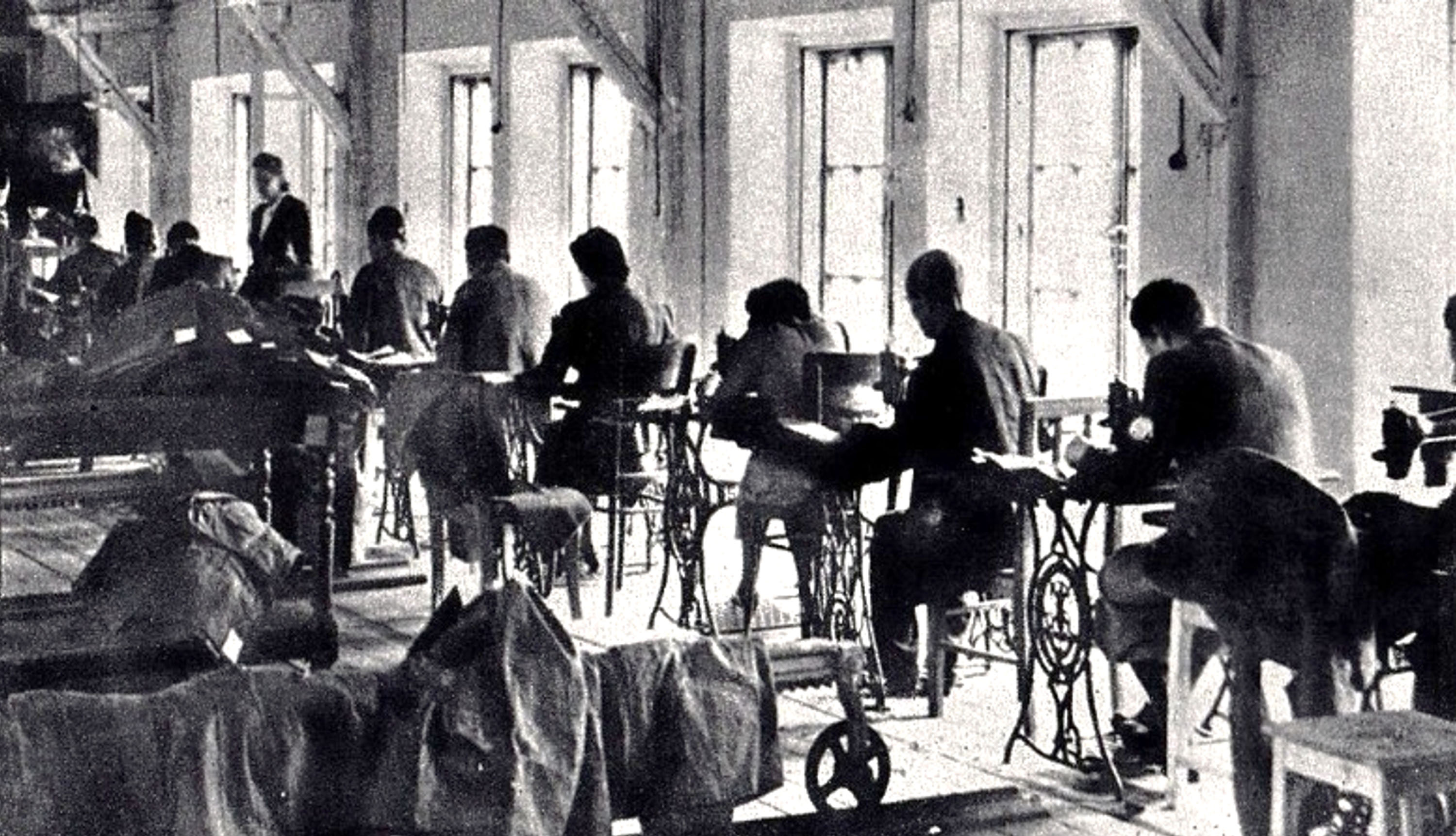 A black-and-white photograph of a workshop with rows of people sitting at sewing machines, working in a long room lit by tall windows.