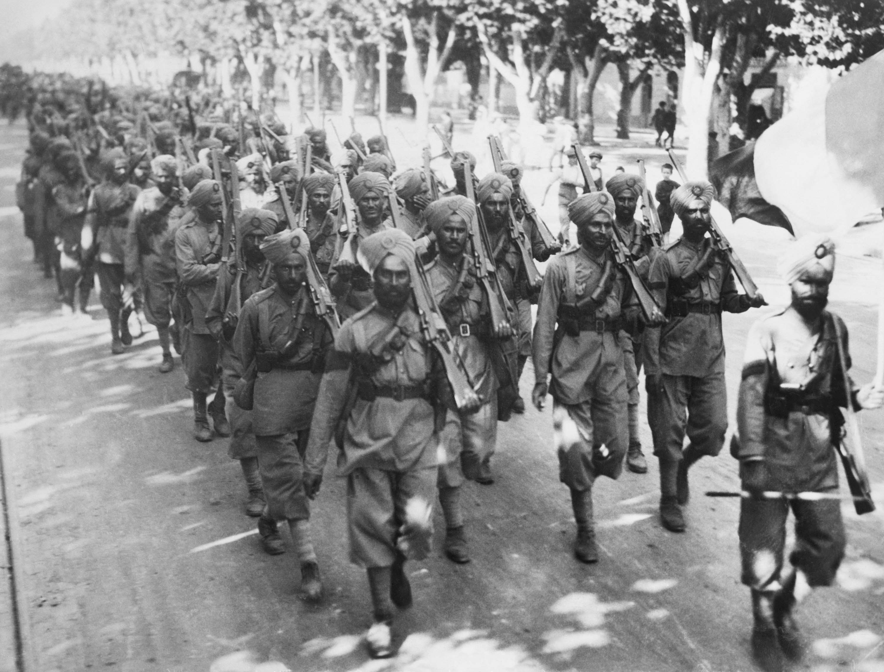 Photo of Indian soldiers marching shoulder-to-shoulder down a paved road, their guns held in each of their left hands.