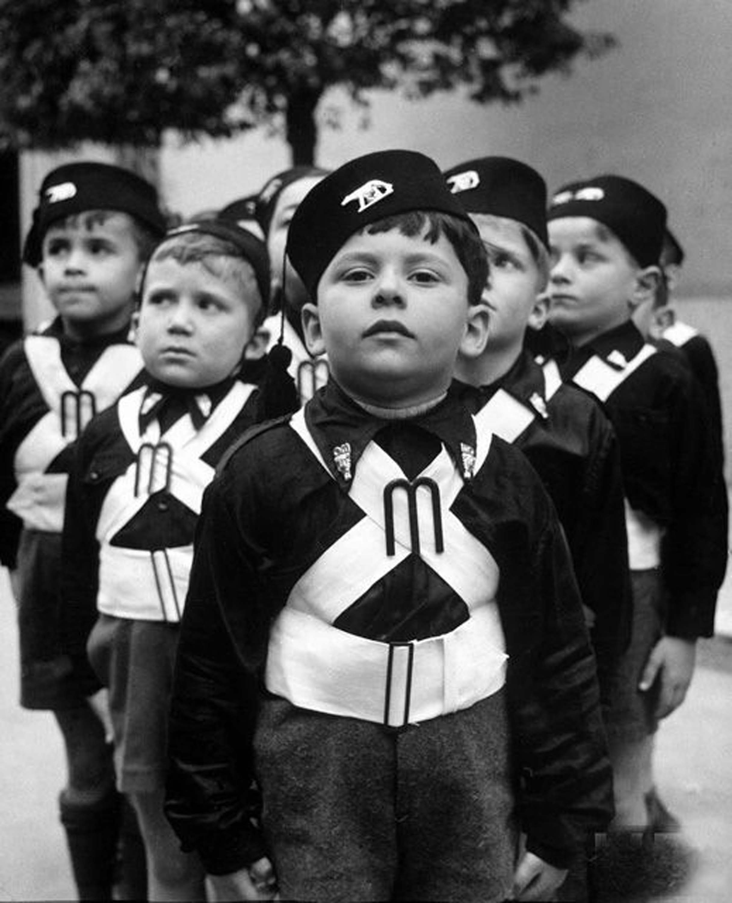 Photo of several young children, dressed in Balilla uniform. They stand with their arms at their sides, wearing long pants and shirts, and white sashes that crisscross their torsos.