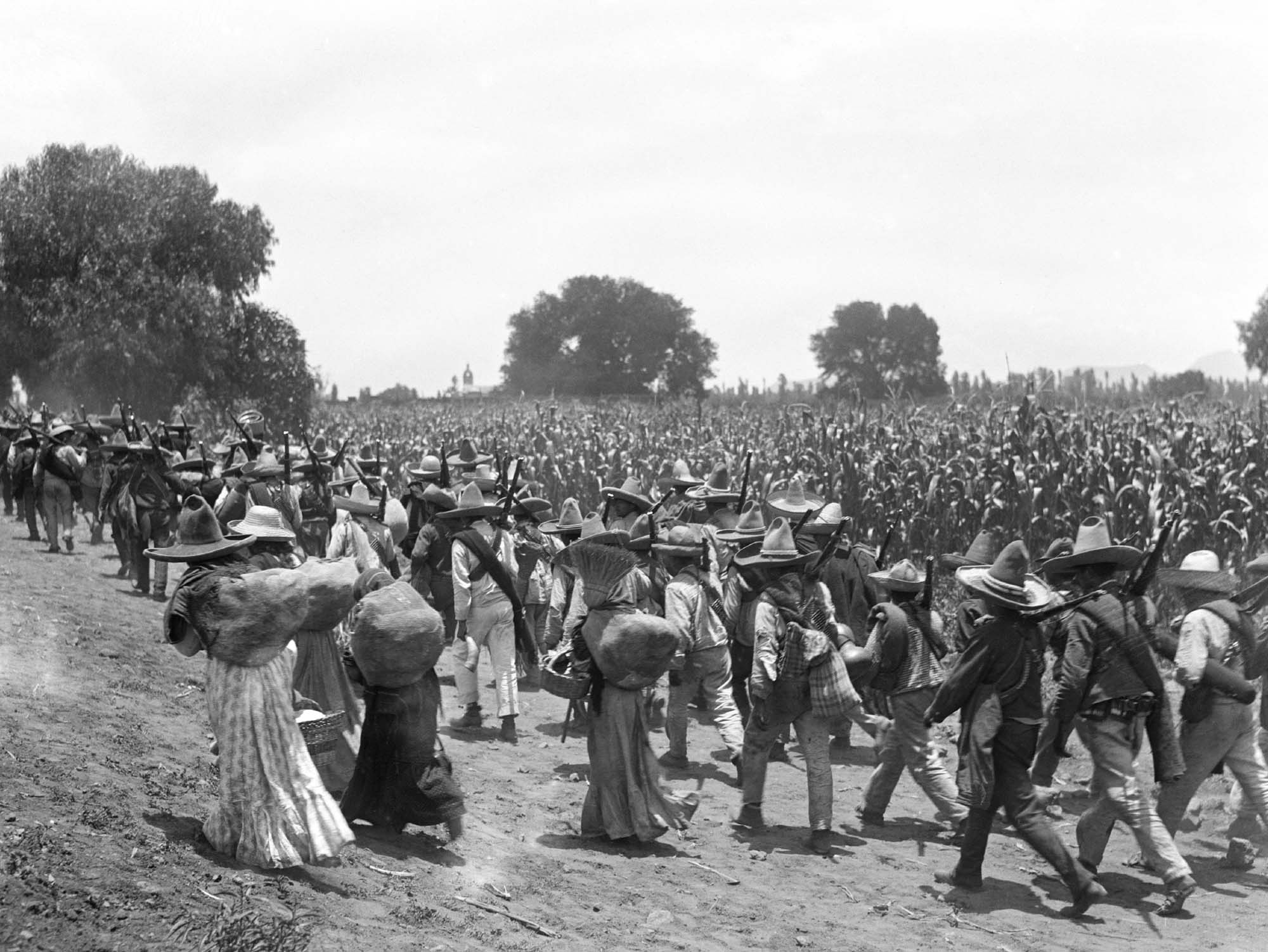 A photograph of a massive group of people marching. Many are holding weapons slung over their shoulders and tall hats to block the sun.