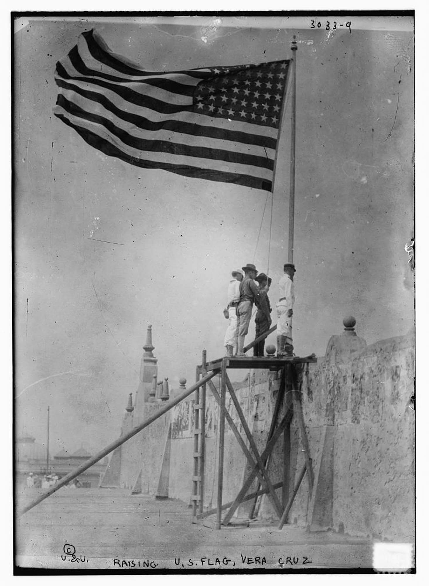 Photo of four men, standing on a raised platform next to a tall stone wall, raising the American flag on a flagpole.