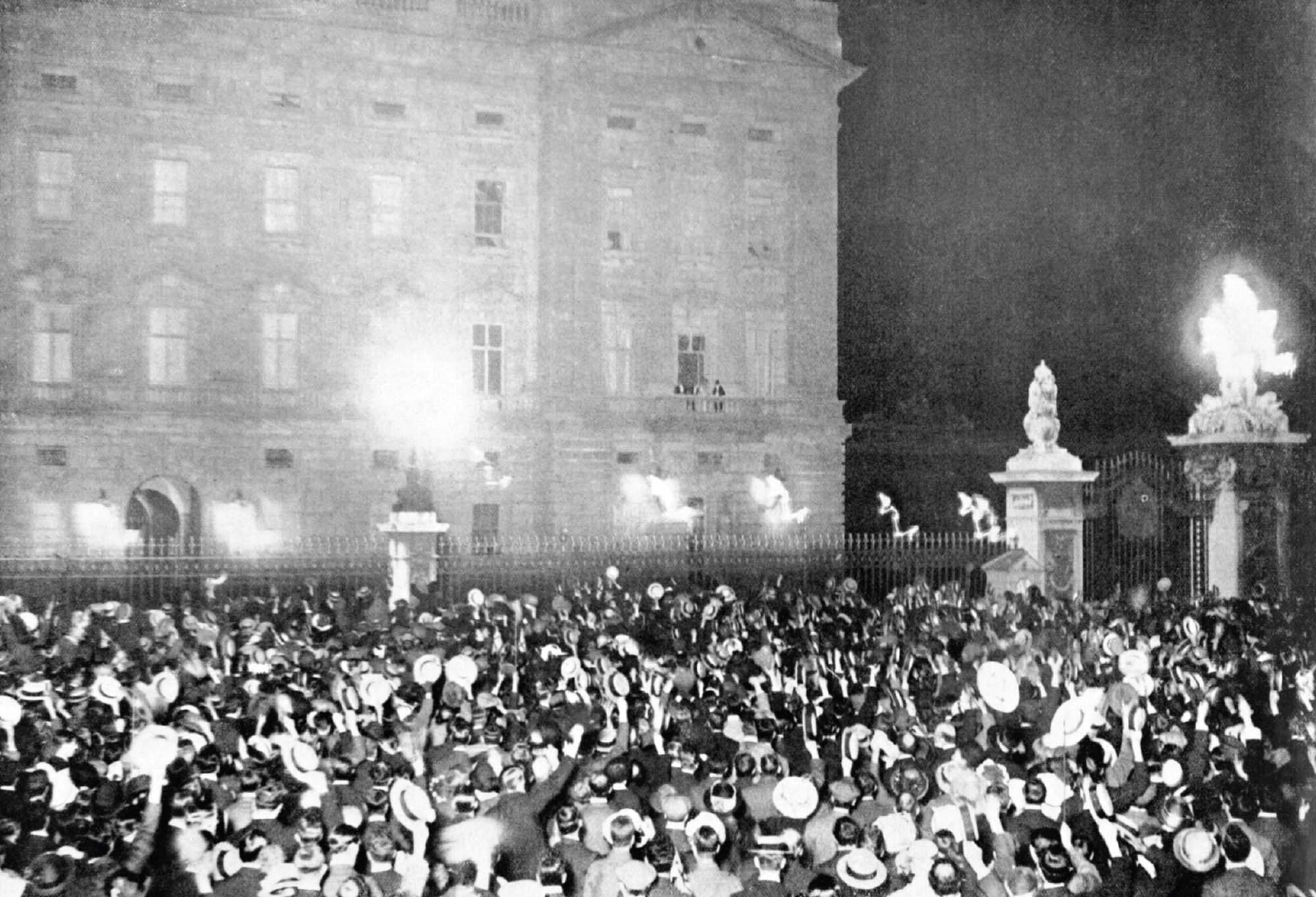 Photo of a large crowd outside of Buckingham Palace waving their arms and hats in the air cheering.