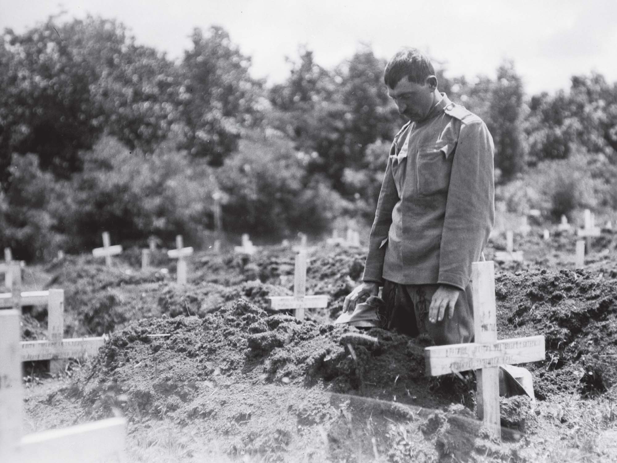 Photo of a man kneeling before a large group of freshly dug graves. Wooden crosses mark each grave site.