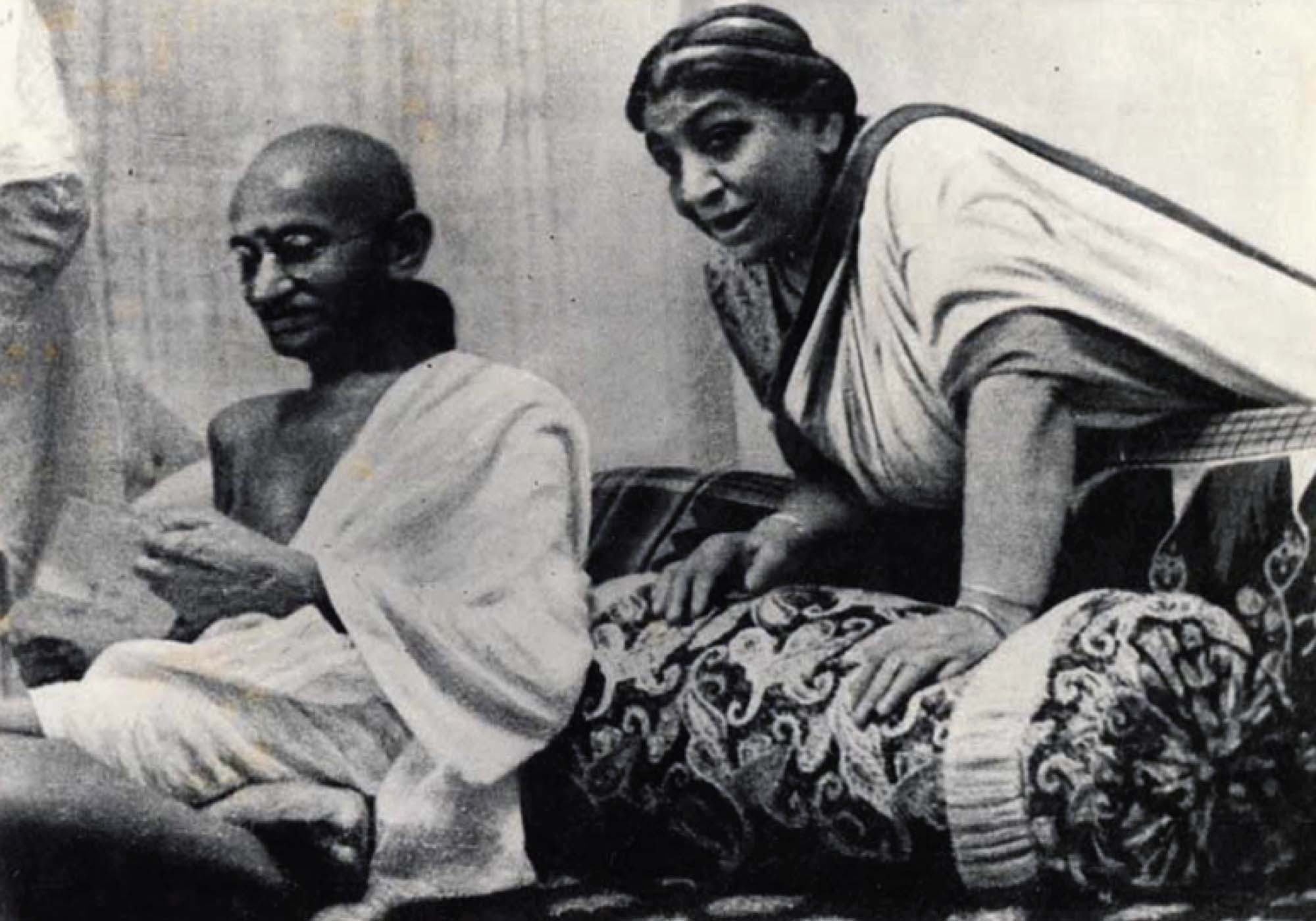 Photo of Gandhi sitting on a couch, flipping through the pages of a pamphlet. Indian activist Sarojini Naidu is leaning over the back of the couch to speak to him.