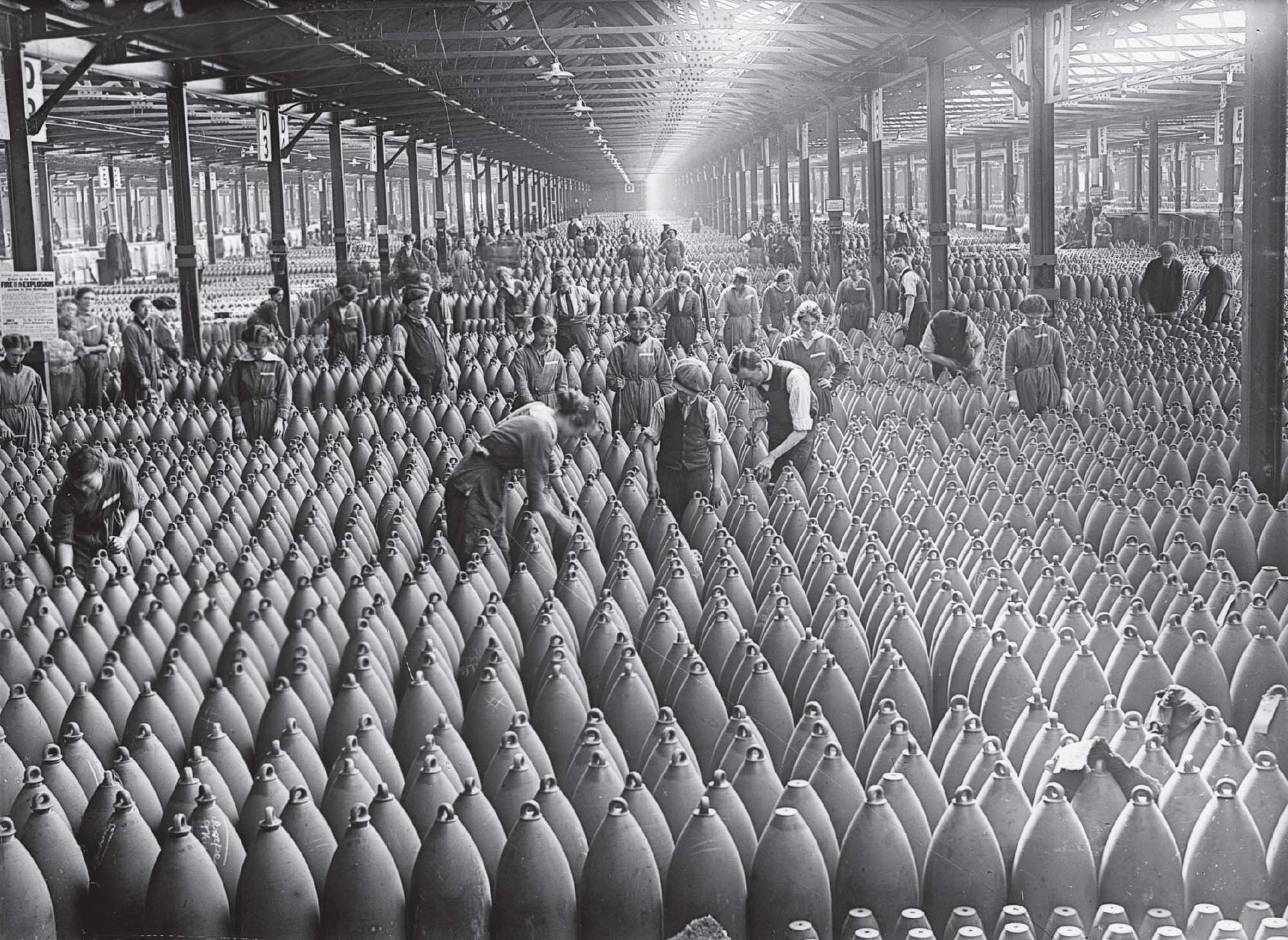 Photograph shows many people standing in a large factory, surrounded by newly made munitions.