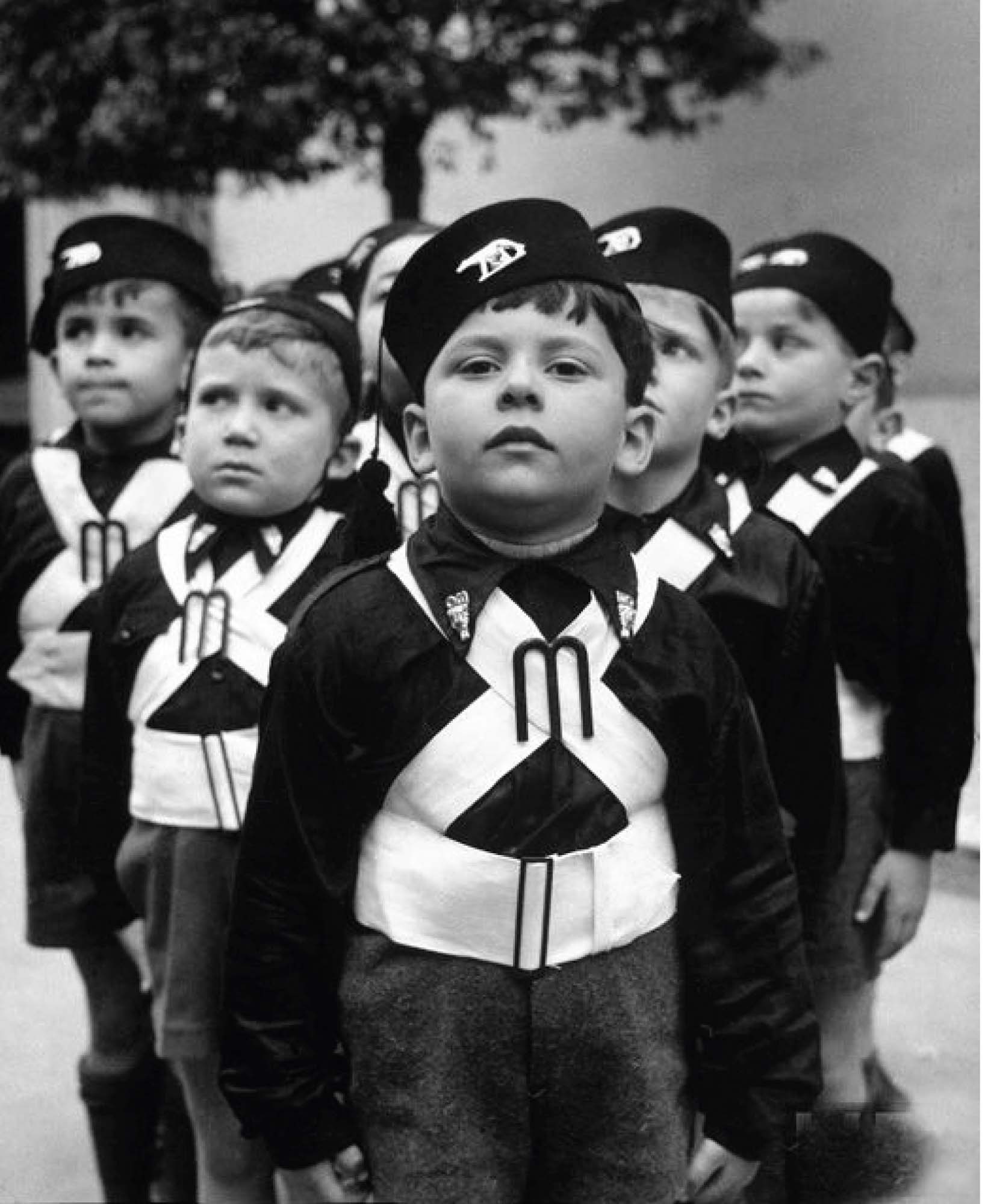 Photo of young Italian boys wearing Ballilla uniforms. These uniforms consist of dark pants, black button up shirts, black hats, and white sashes that criss-cross over the boys’ bodies.