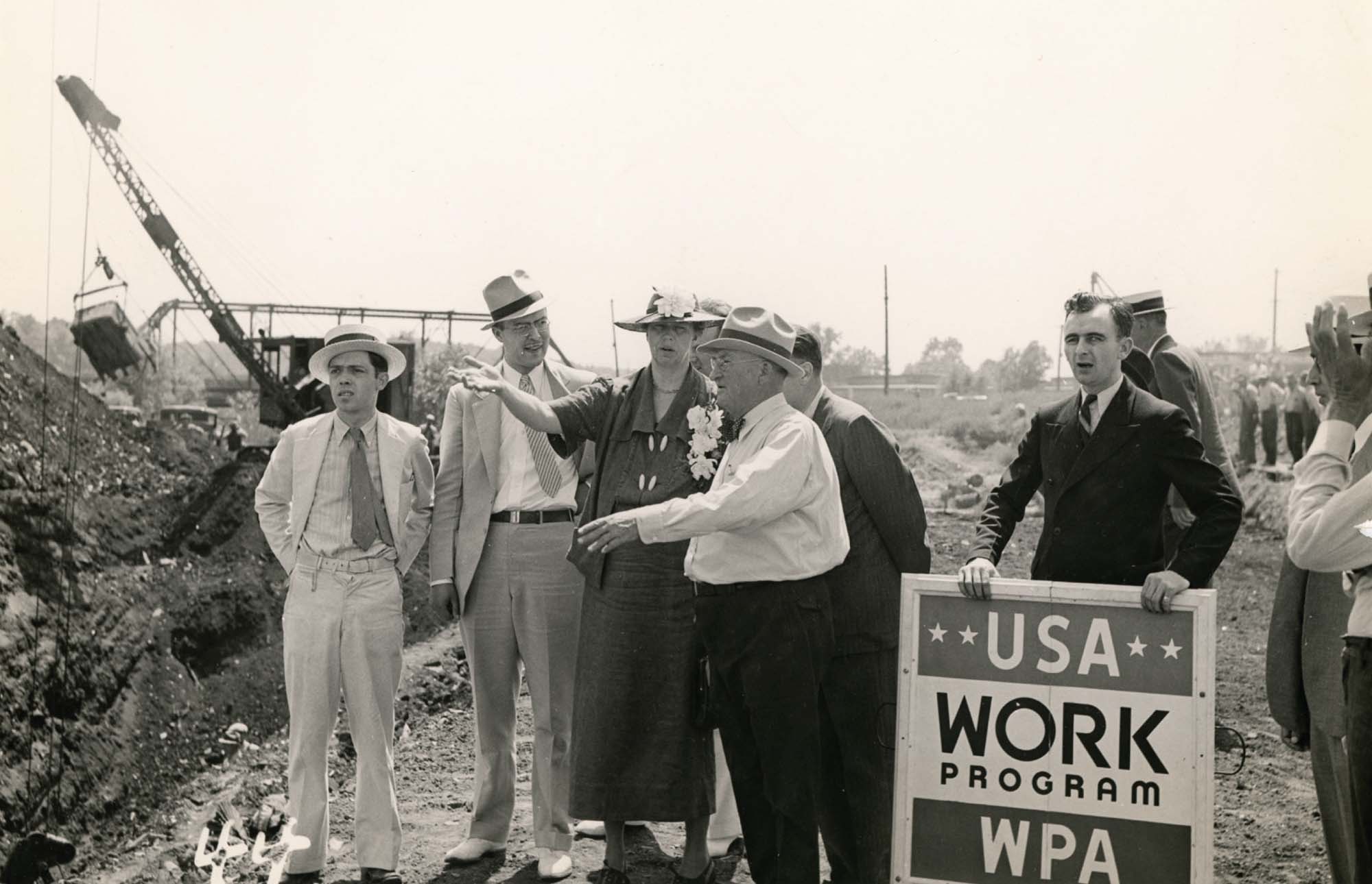 Photo of Elanor Roosevelt and several men standing outside at a construction site. One man is holding a sign that reads “USA Work Program”.
