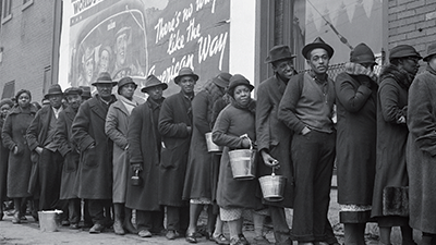 A photograph shows African-American flood victims lined up to get food & clothing from Red Cross relief station in front of billboard extolling, ironically, WORLD’S HIGHEST STANDARD OF LIVING/ THERE’S NO WAY LIKE THE AMERICAN WAY.