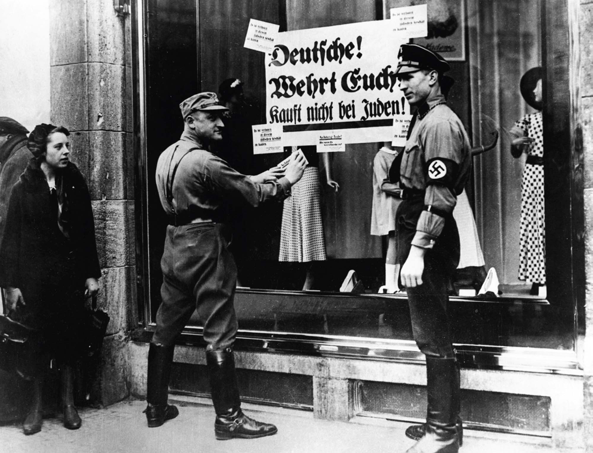 Photo of Nazis placing a sign on the window of Jewish business. The sign reads, “Germans! Defend Yourselves! Don’t buy from Jews!”