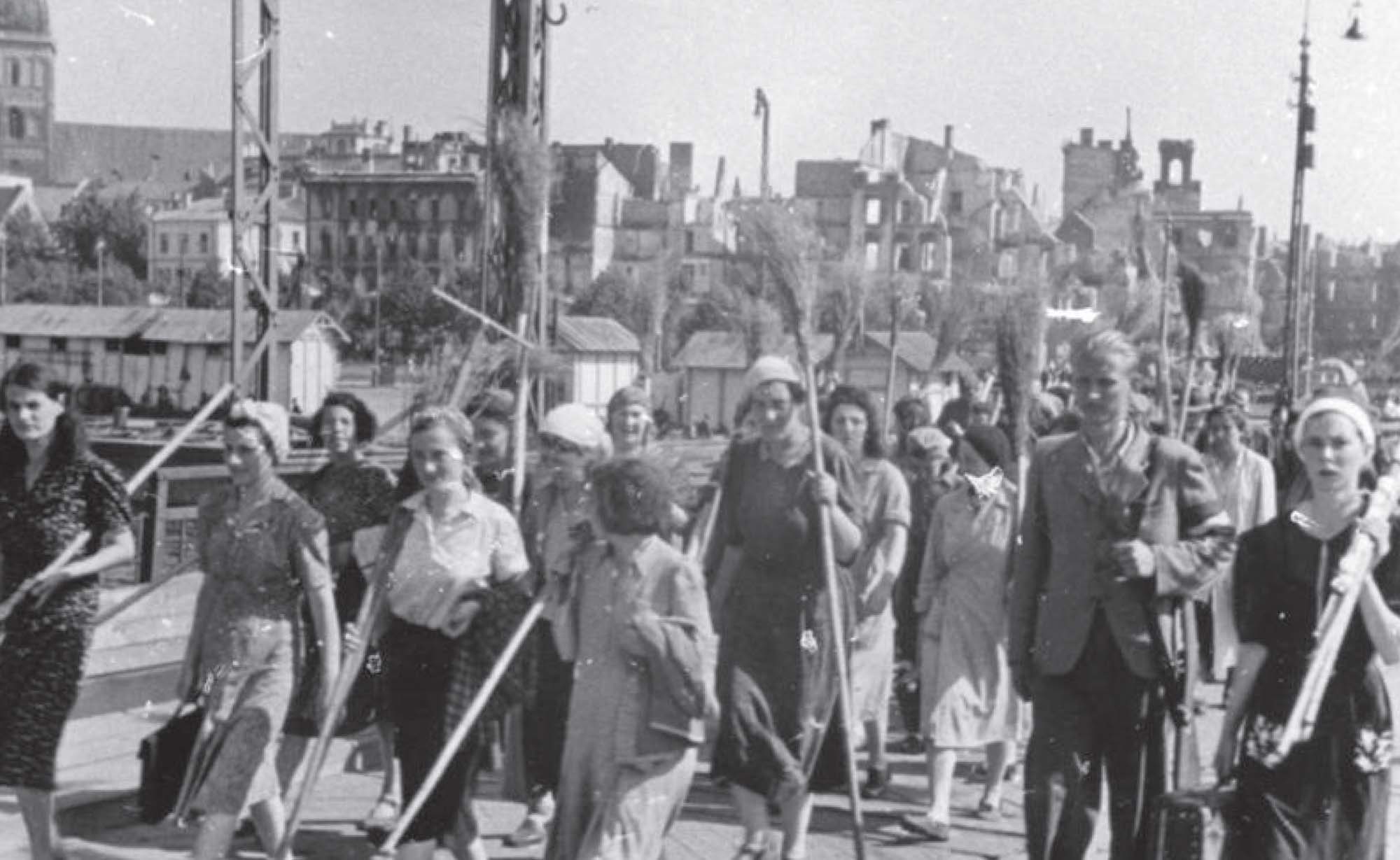 A black and white photograph of many people, marching down the street holding brooms in their hands. Behind them are several large buildings.