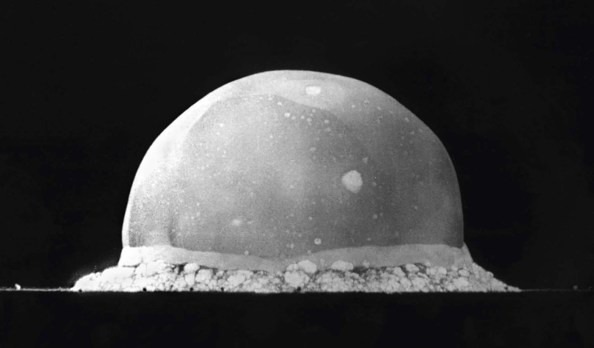 Photograph shows a fire ball just after detonation, which is dome shaped and balanced on a pile of, what looks like, dust.