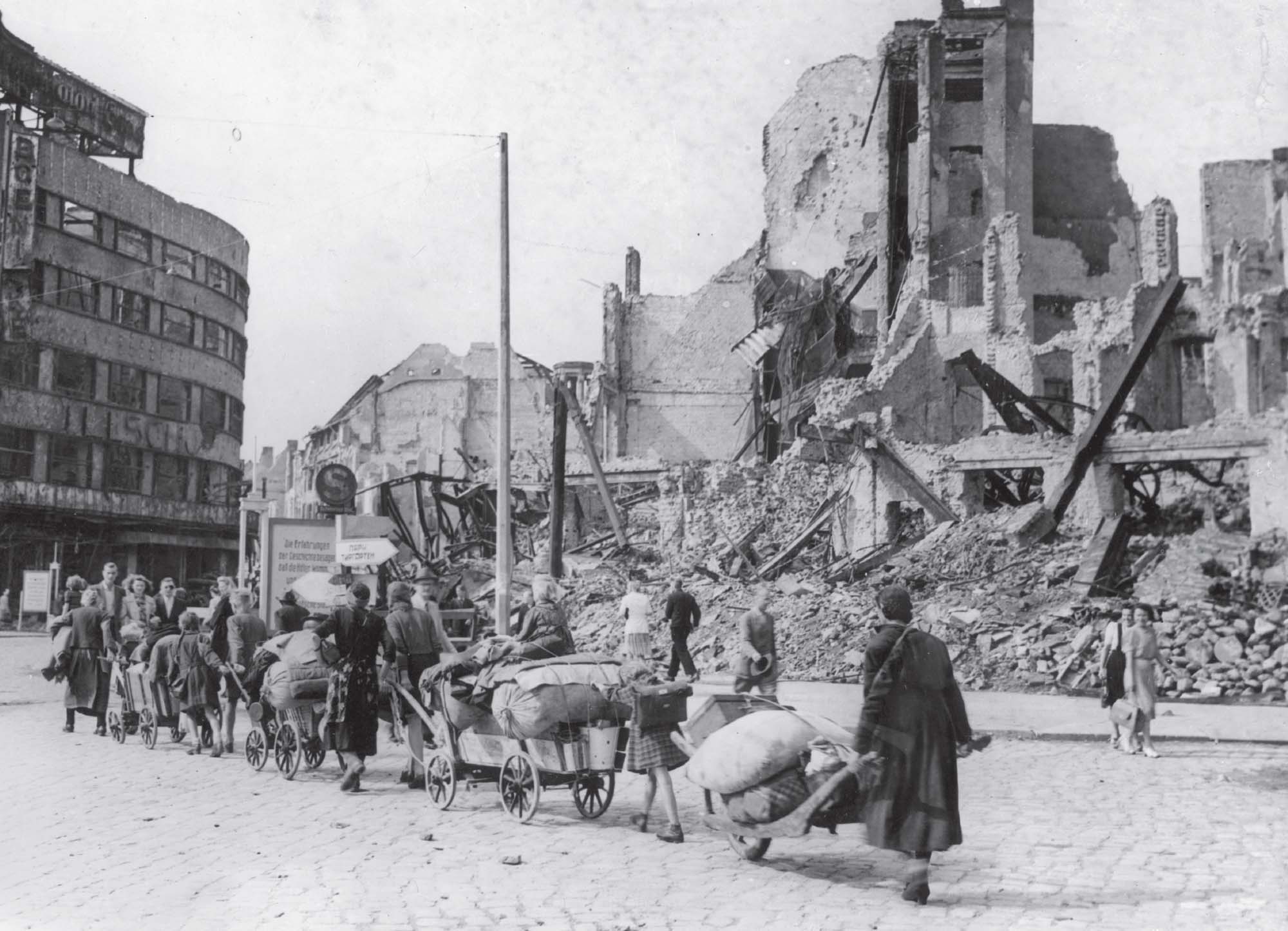 Photo shows a caravan of people walking together, hauling belongings on wagons, next to a destroyed building and piles of rubble.