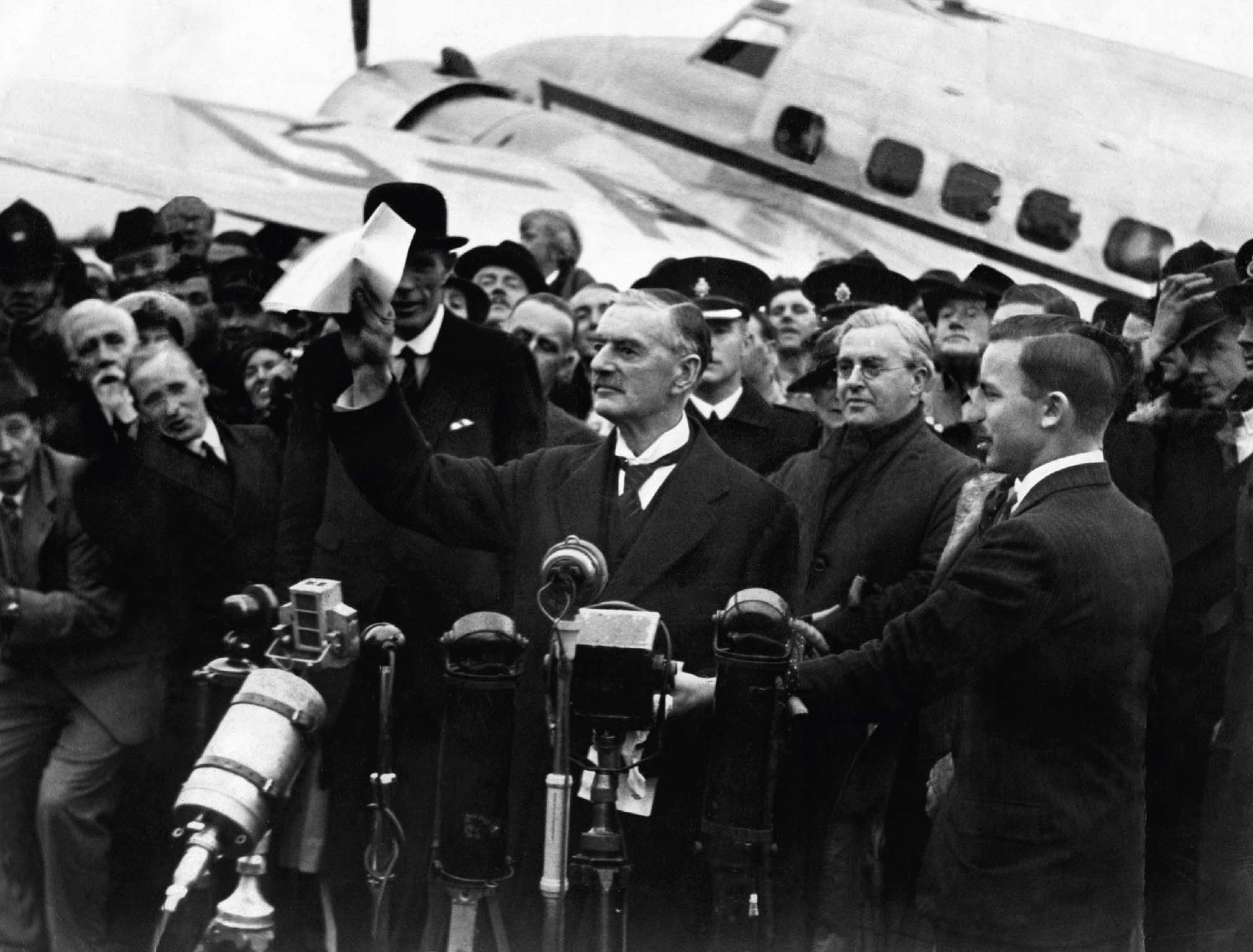 Photo of British Prime Minister Neville Chamberlin standing in front of a set of microphones and holding a piece of paper up in the air. A large crowd is gathered behind him.