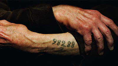 A close-up photograph of the forearms of a Holocaust survivor. There is a number tattooed on one of the person’s forearms.