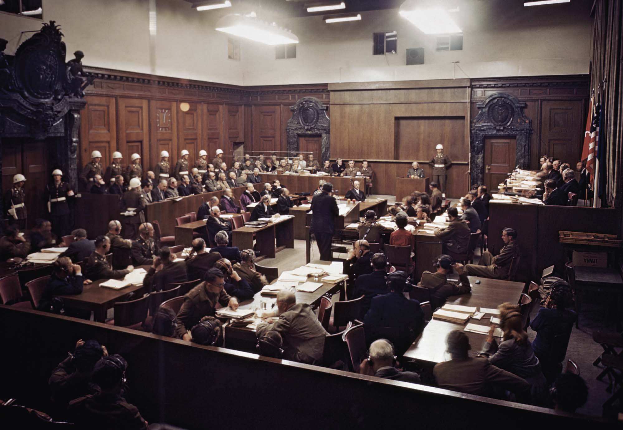 Photo of a crowded courtroom at the Nuremberg trials. Those in attendance can be seen wearing headsets so that they were able to listen to the proceedings in their own native language.