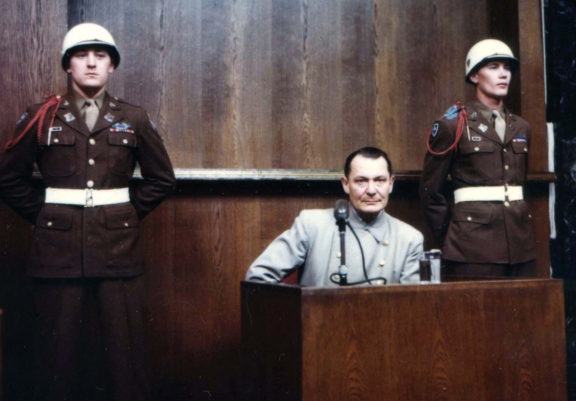 Photo of Hermann Goering sitting behind a witness stand at his trial in Nuremberg. Two military guards stand behind him.