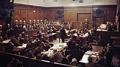 Photo of a crowded courtroom at the Nuremberg trials. Those in attendance can be seen wearing headsets so that they were able to listen to the proceedings in their own native language.