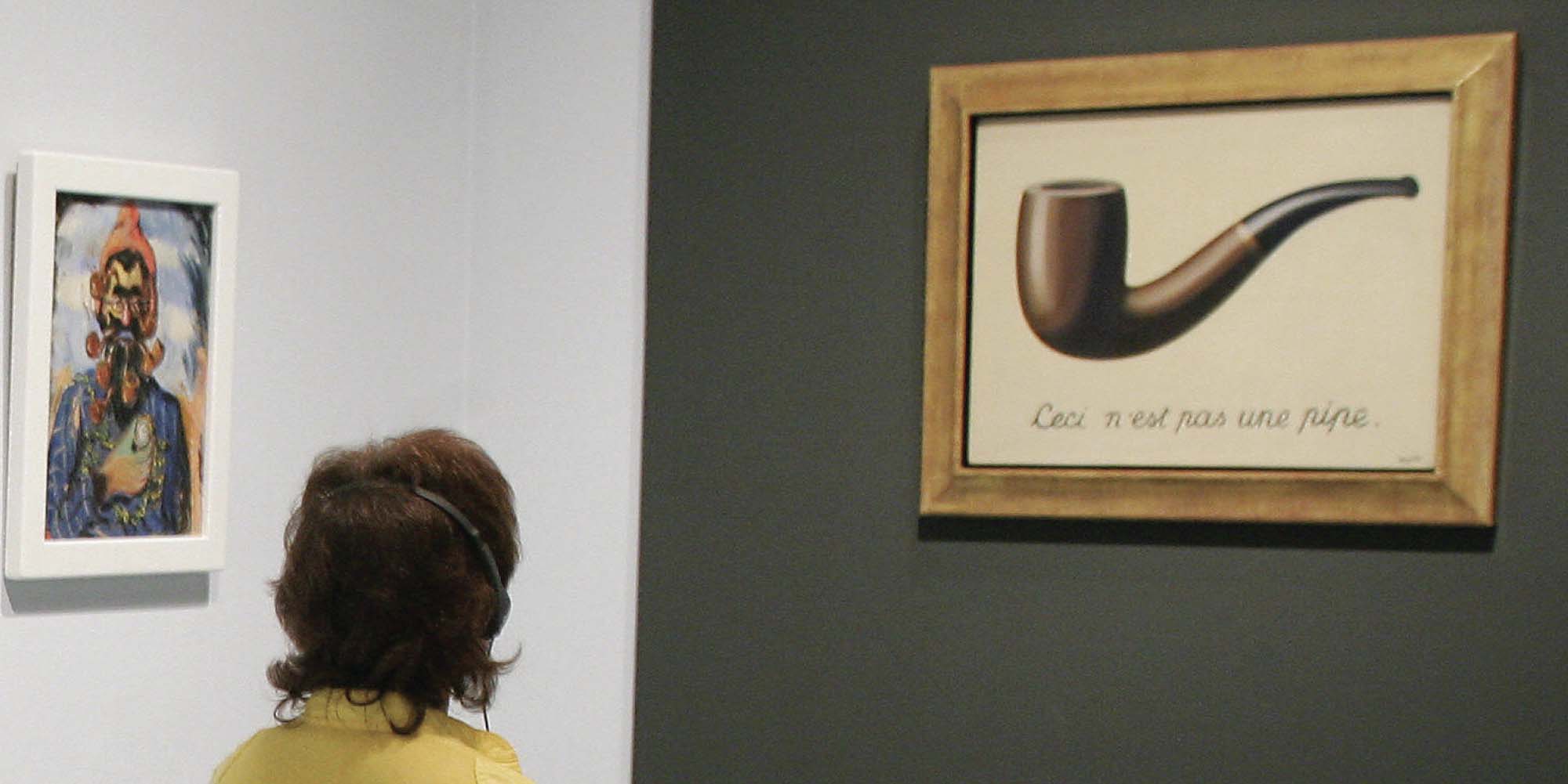 A museum visitor looks at a paiting of of a wooden pipe with black text below that reads, “Ceci n’est pas une pipe”.