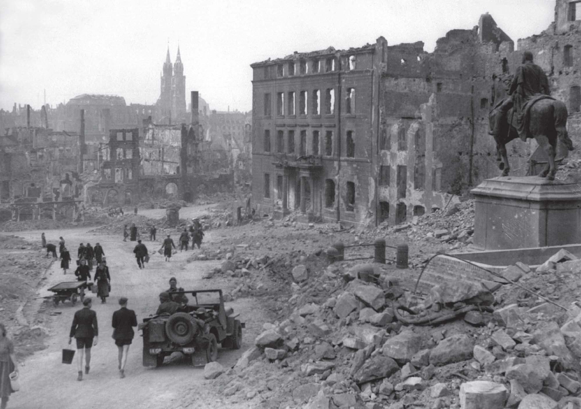 People walk through a city that has been reduced to ruins. A statue of a man on horseback stands on top of a pile of rubble, next to a bombed-out building. One person drives a military vehicle through the wreckage and several others walk through.  